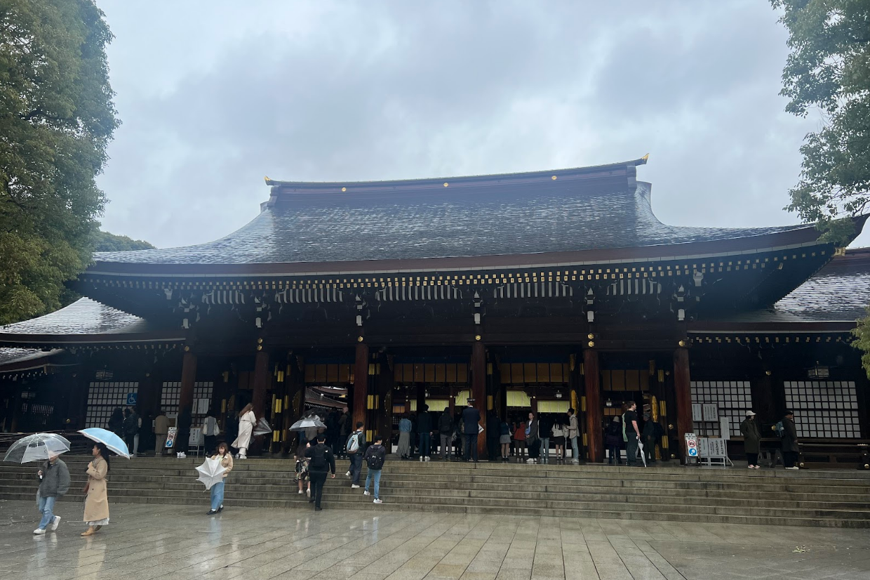An image of people walking up the steps into a large temple with dark features. There are trees and rain clouds in the surrounding view.