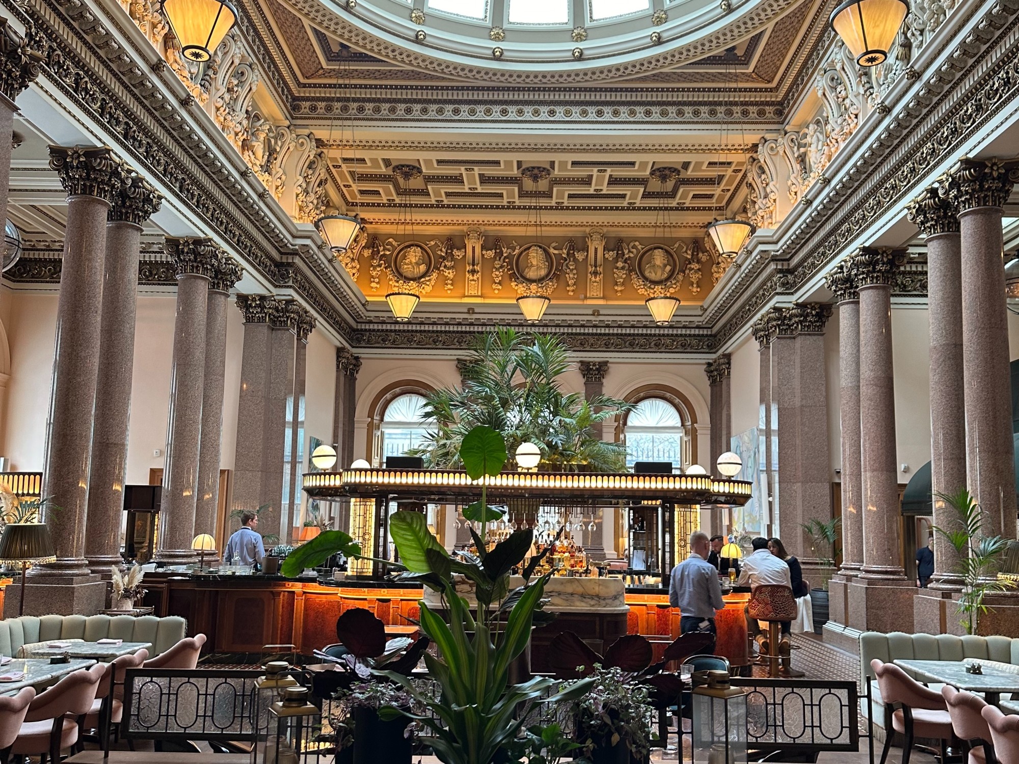 A luxury dining room and bar of a hotel, with ancient Greek-style columns, sculptures in the recessed ceiling, floral arrangements, and soft, amber lighting fixtures.