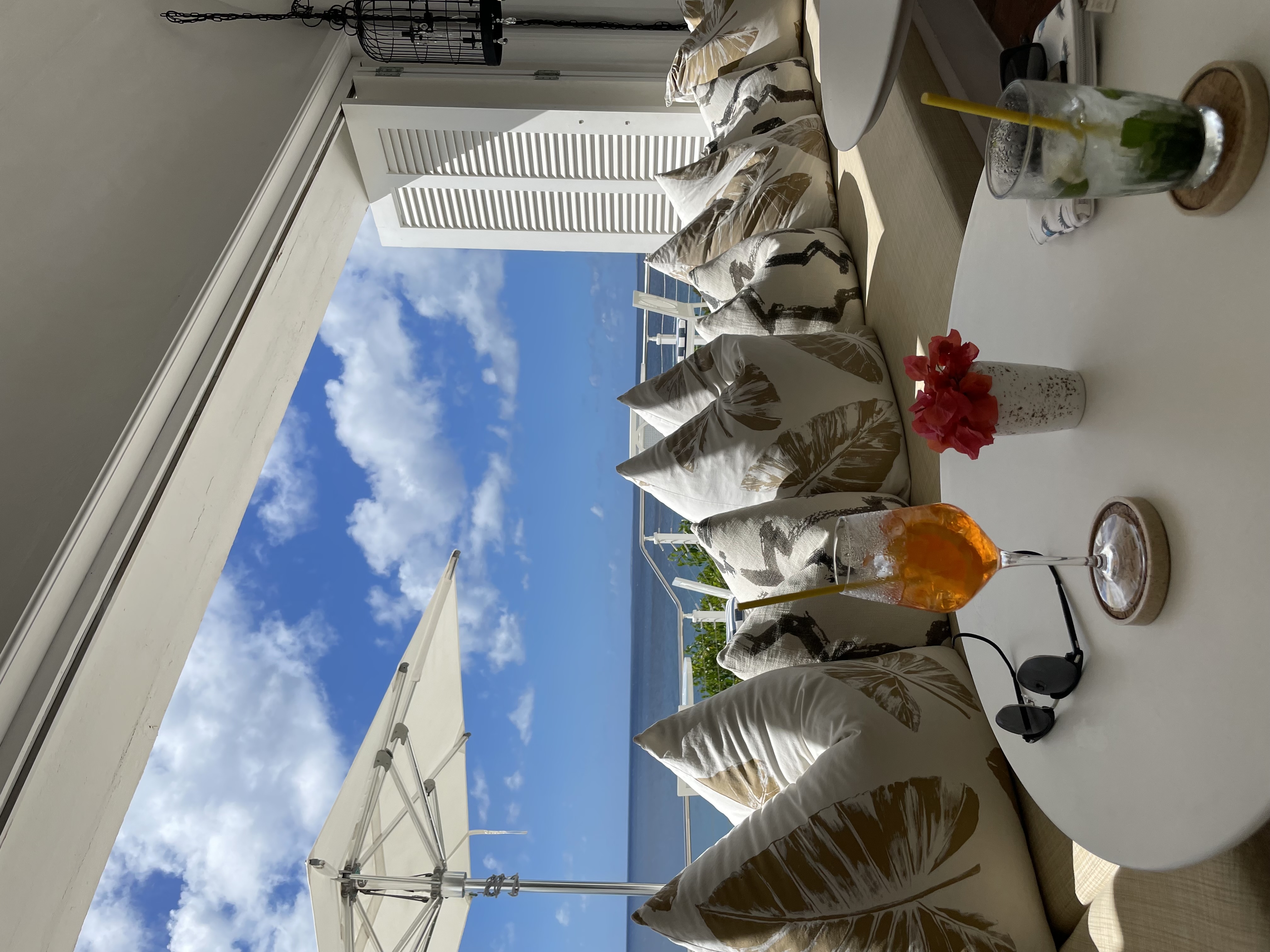 Drinks on a white table with a row of white patterned cushions behind by a large window with white shutters with ocean view