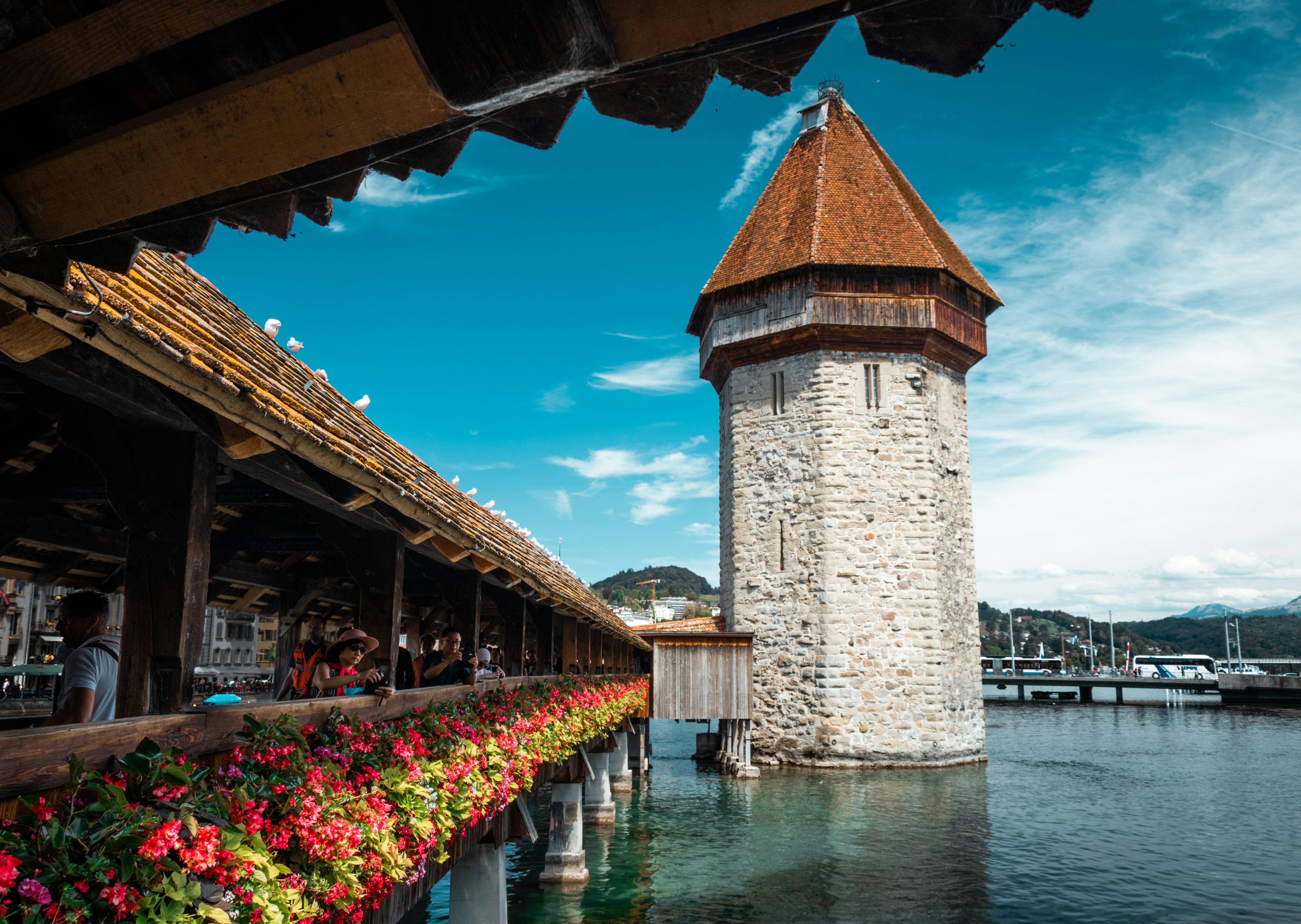 The image depicts a charming European scene featuring a stone tower with a conical roof beside a flower-adorned covered wooden bridge, overlooking a tranquil lake and clear skies.