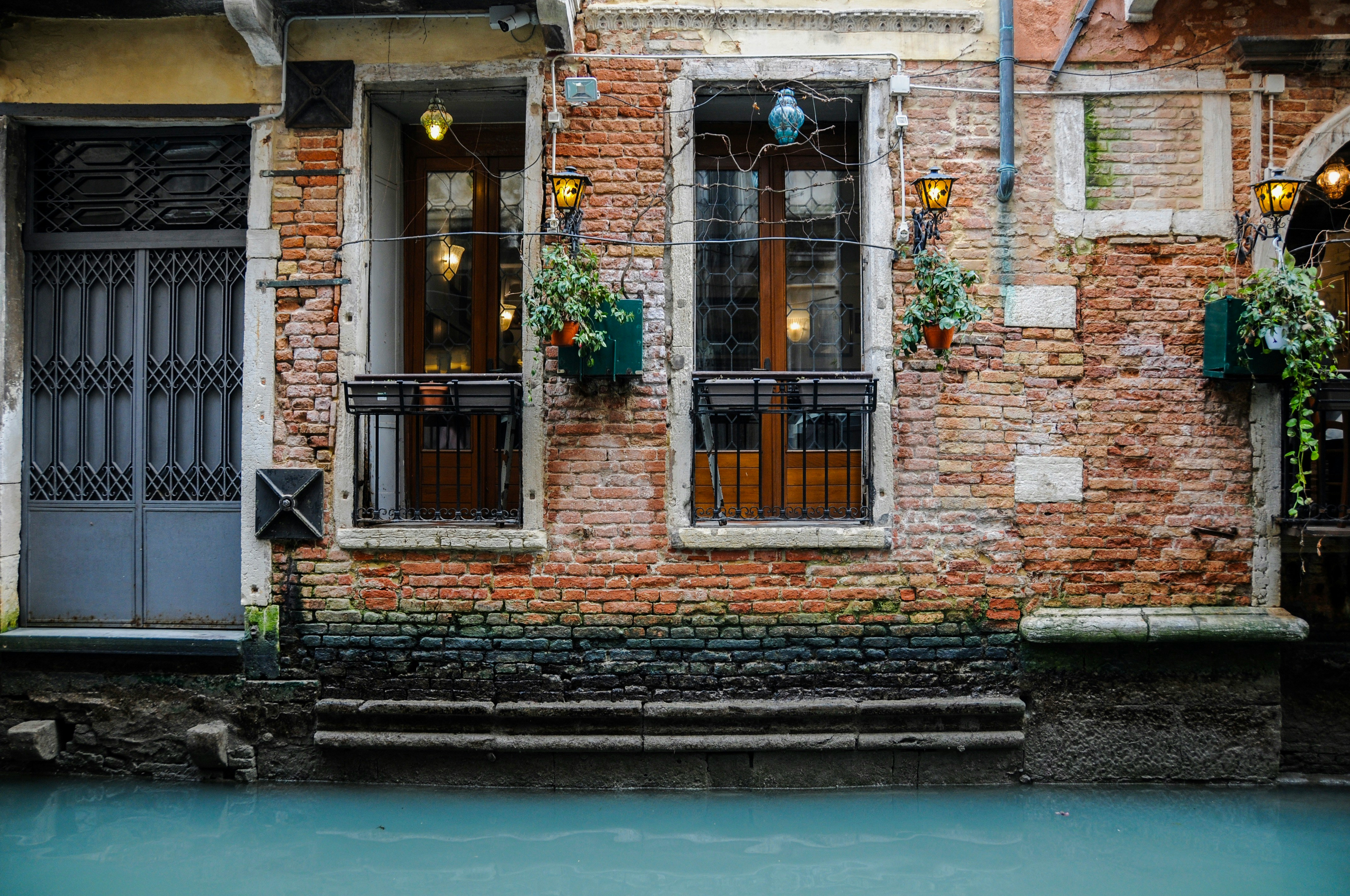A brown brick building next to a canal during the daytime. There are potted plants and balconies near the window.
