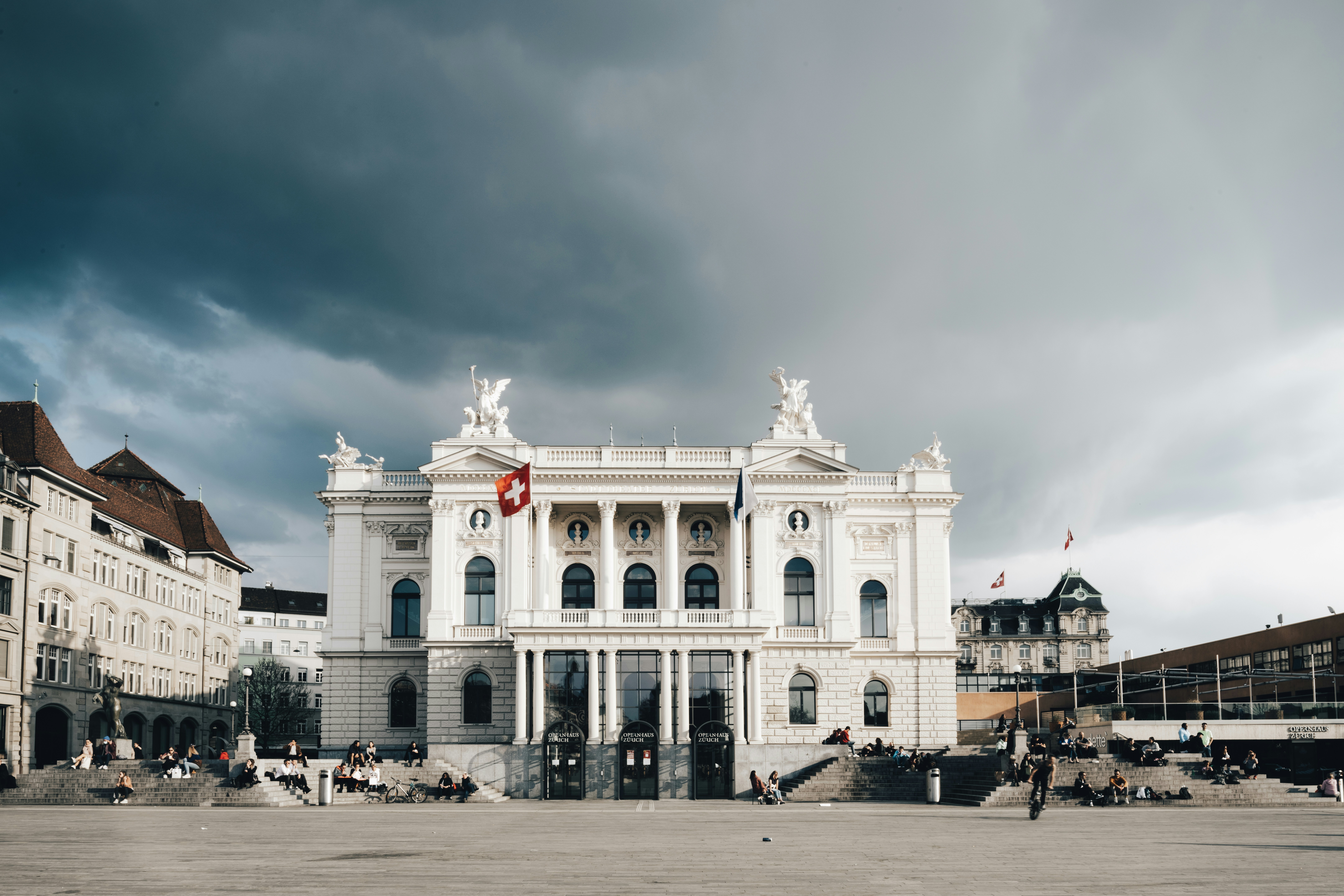 A picture of people walking near a white concrete building with a Swiss flag during daytime.
