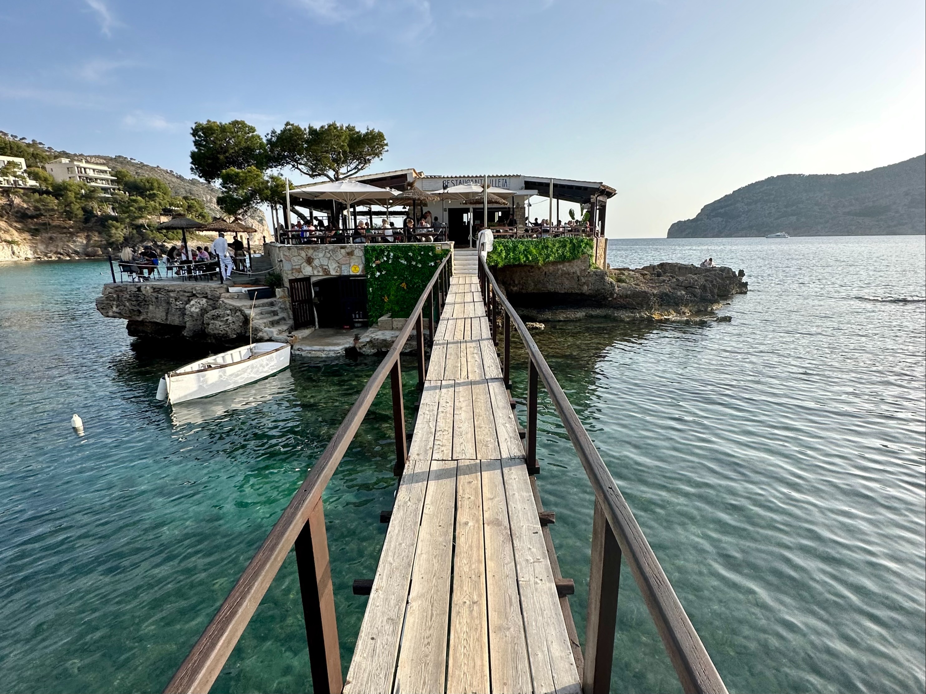 A wooden boardwalk surrounded by water with a venue in the distance beneath trees and cabanas.