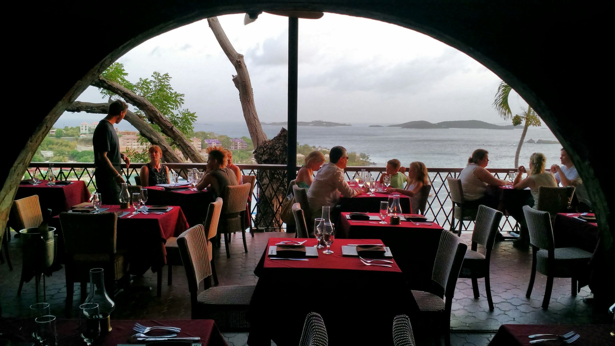 A view of a restaurant with red tablecloths, an arched ceiling, people dining and a view of the sea and trees in the surrounding areas.