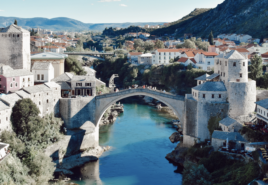 A view of a blue river beneath a tall, stone arched bridge and various old stone buildings in the surrounding area. There is also a mountain on the right.