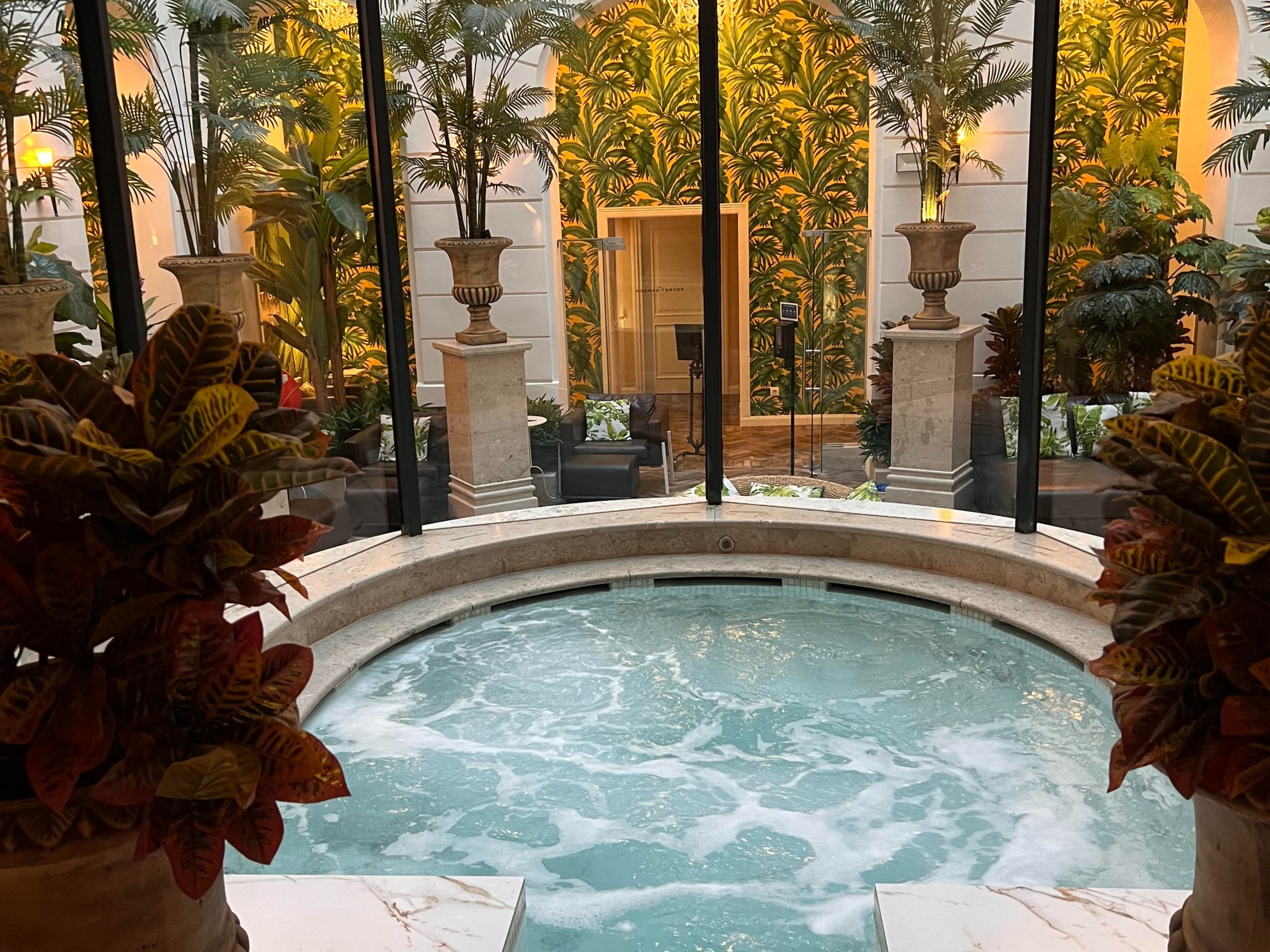 A hot thermal pool inside of a hotel. There are palm leaves, patterned wall paper and lounge chairs in the background.