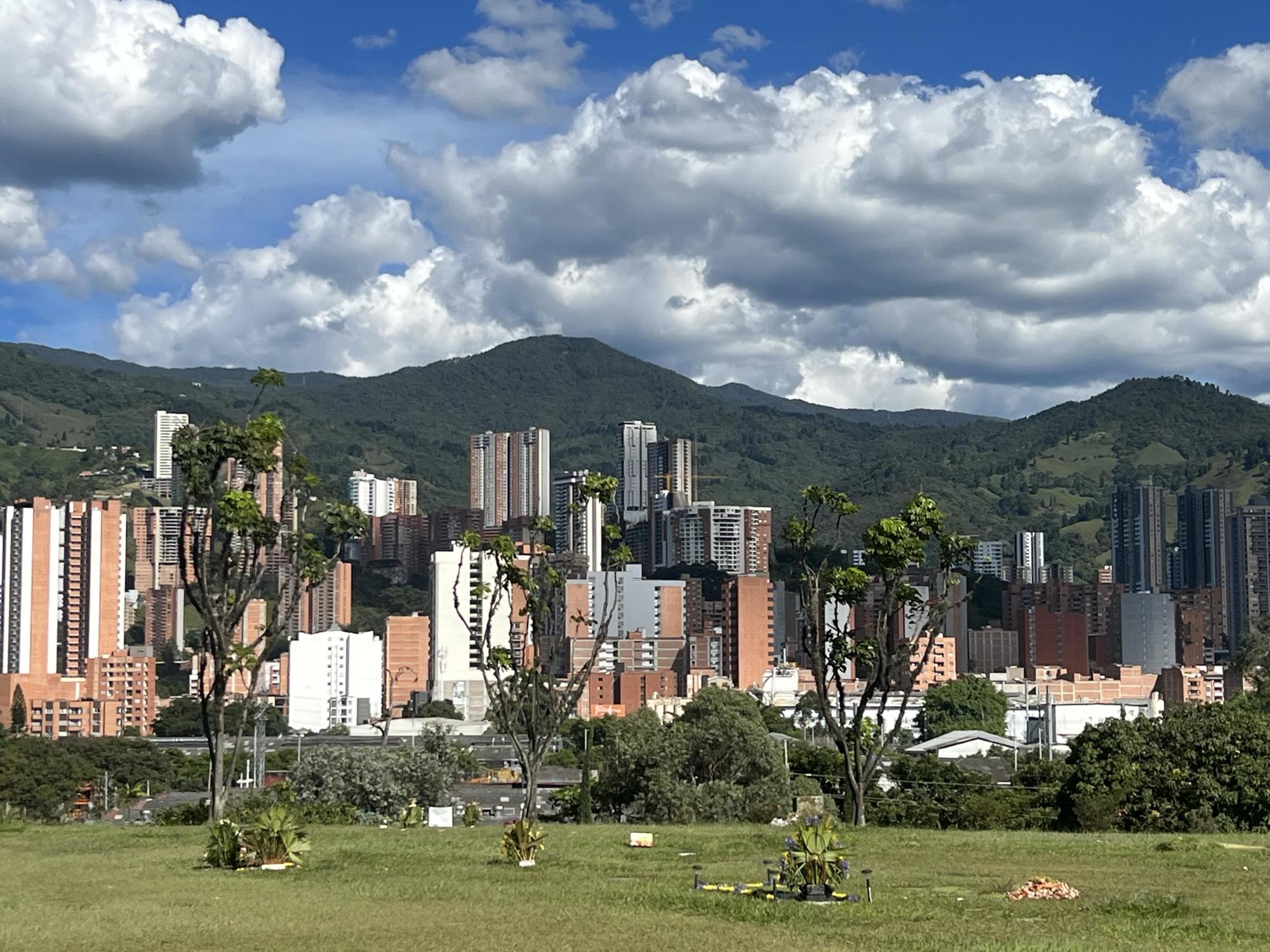 A city skyline against green mountains beneath a cloudy blue sky.