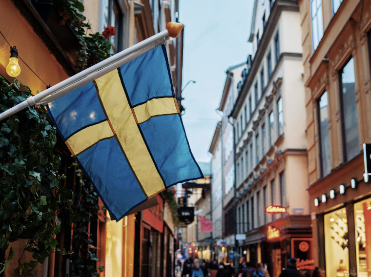 A Swedish flag hanging from a building and people walking in an alleyway in the background.