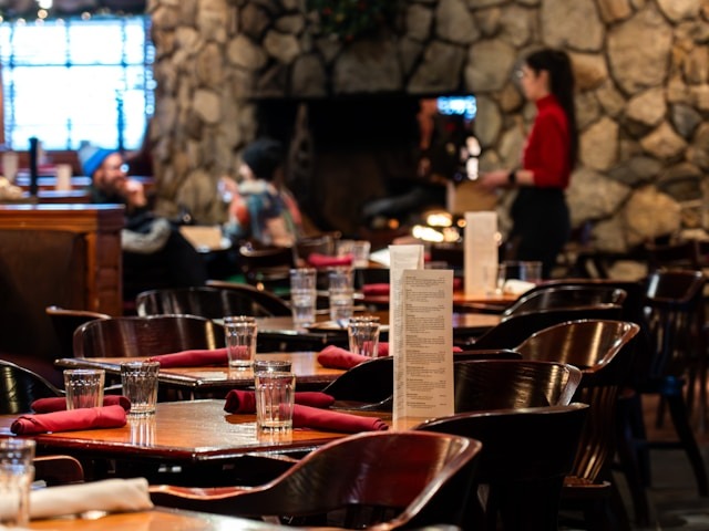 A restaurant complete with wooden tables and chairs and a stone walled fireplace in the background.