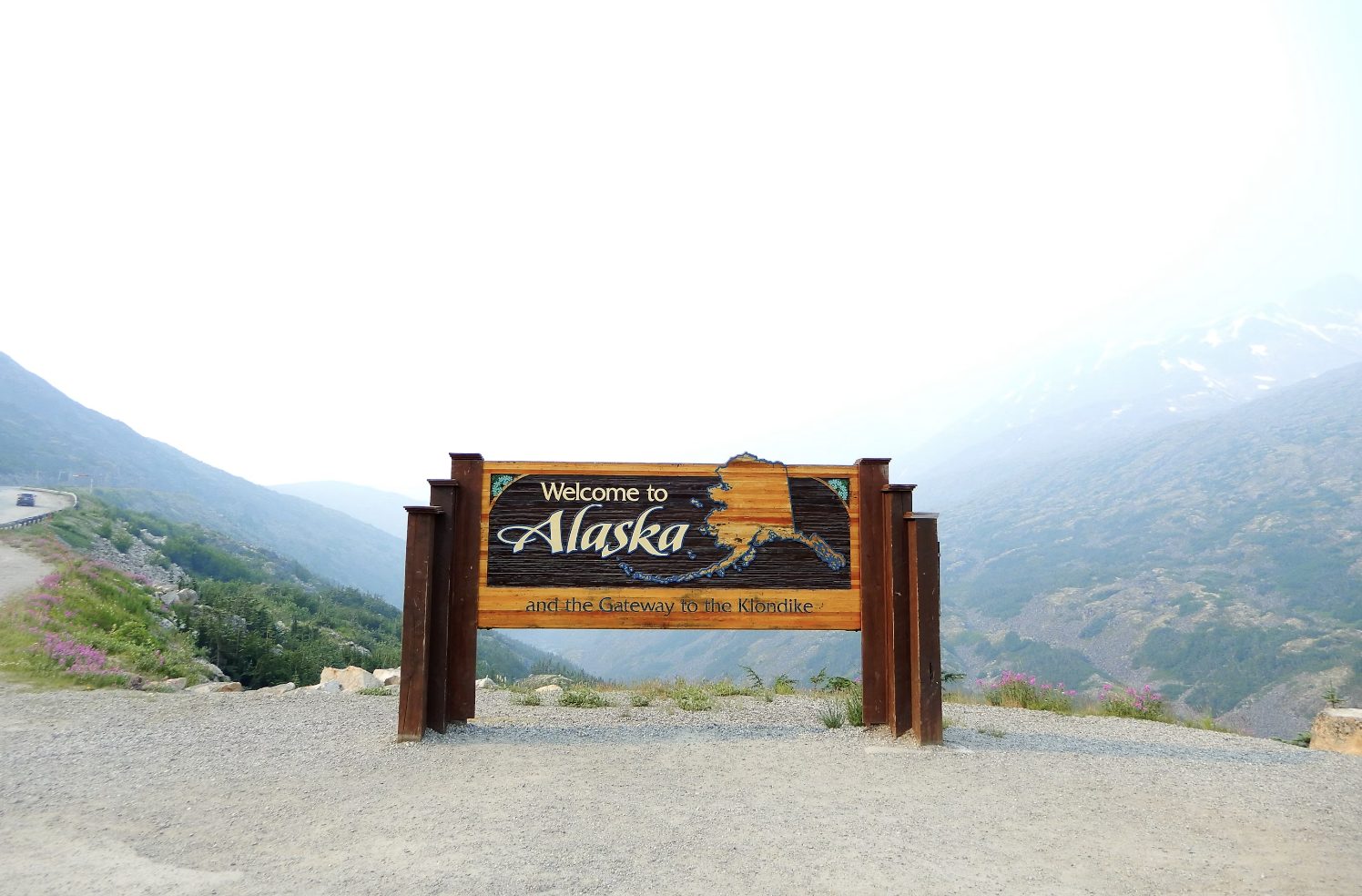 A welcome to Alaska sign on the side of the road with mountains in the background.