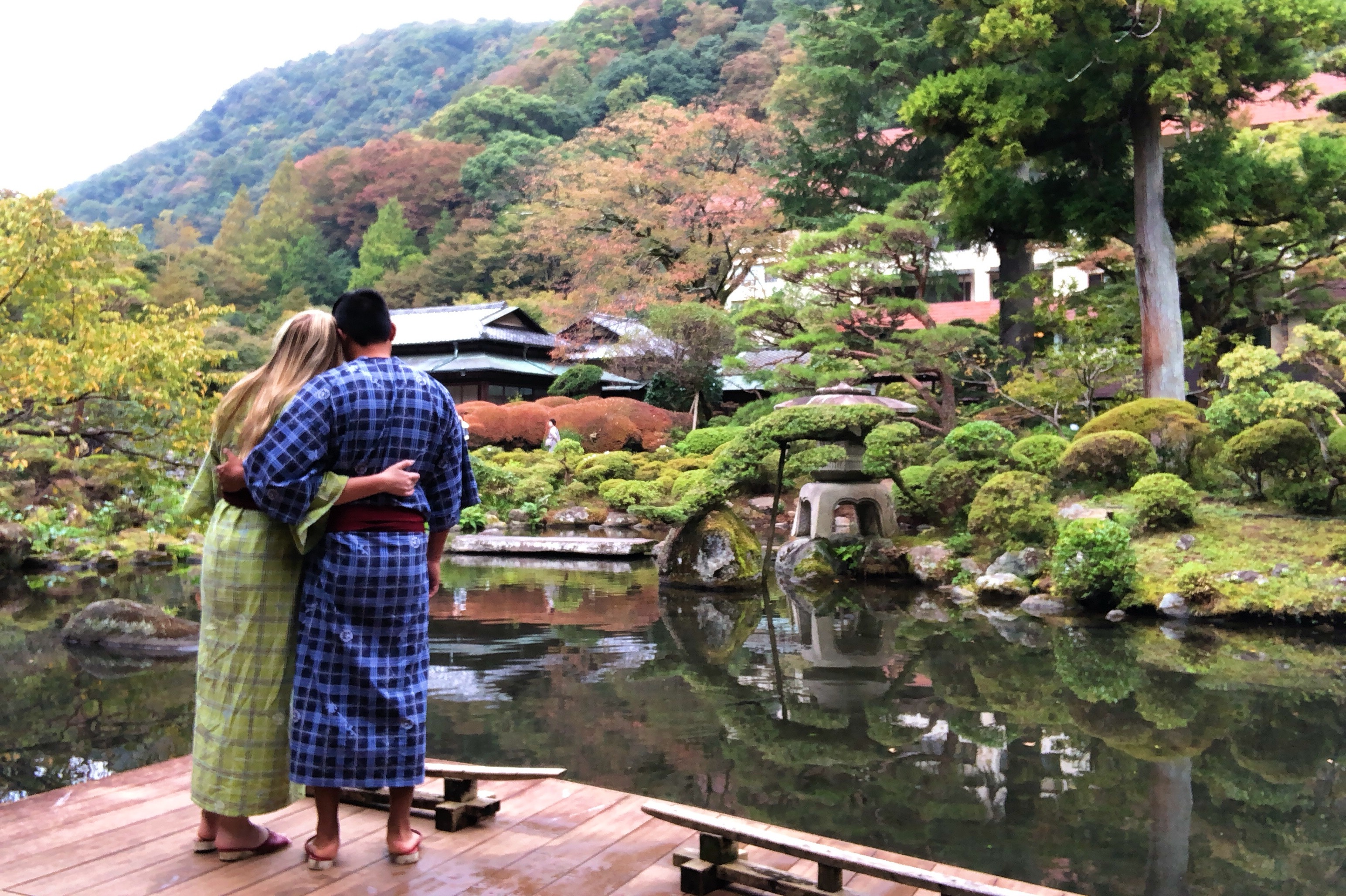 A couple wearing robes and posing for a photo in front of a pond, forest and houses.