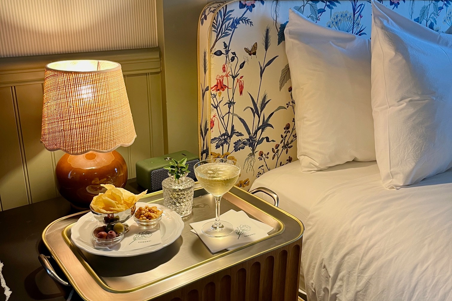 A view of a hotel bedroom complete with white bedding, a floral patterned headboard, side table lamp turned on and tray of food and a cocktail.