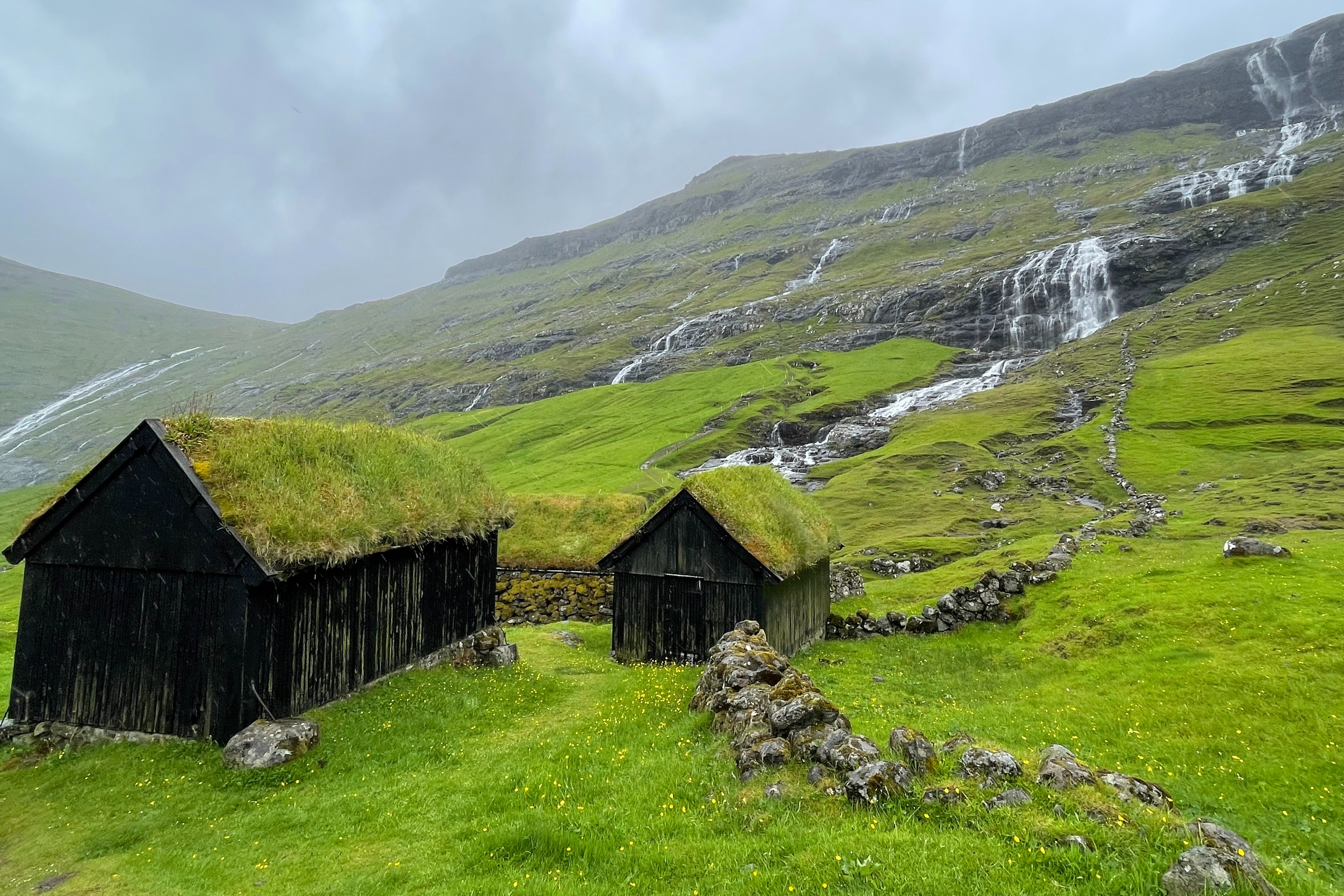 A picturesque village nestled in a dramatic valley with grass covered roofs on top of wooden shacks and water cascading down a grassy hill.