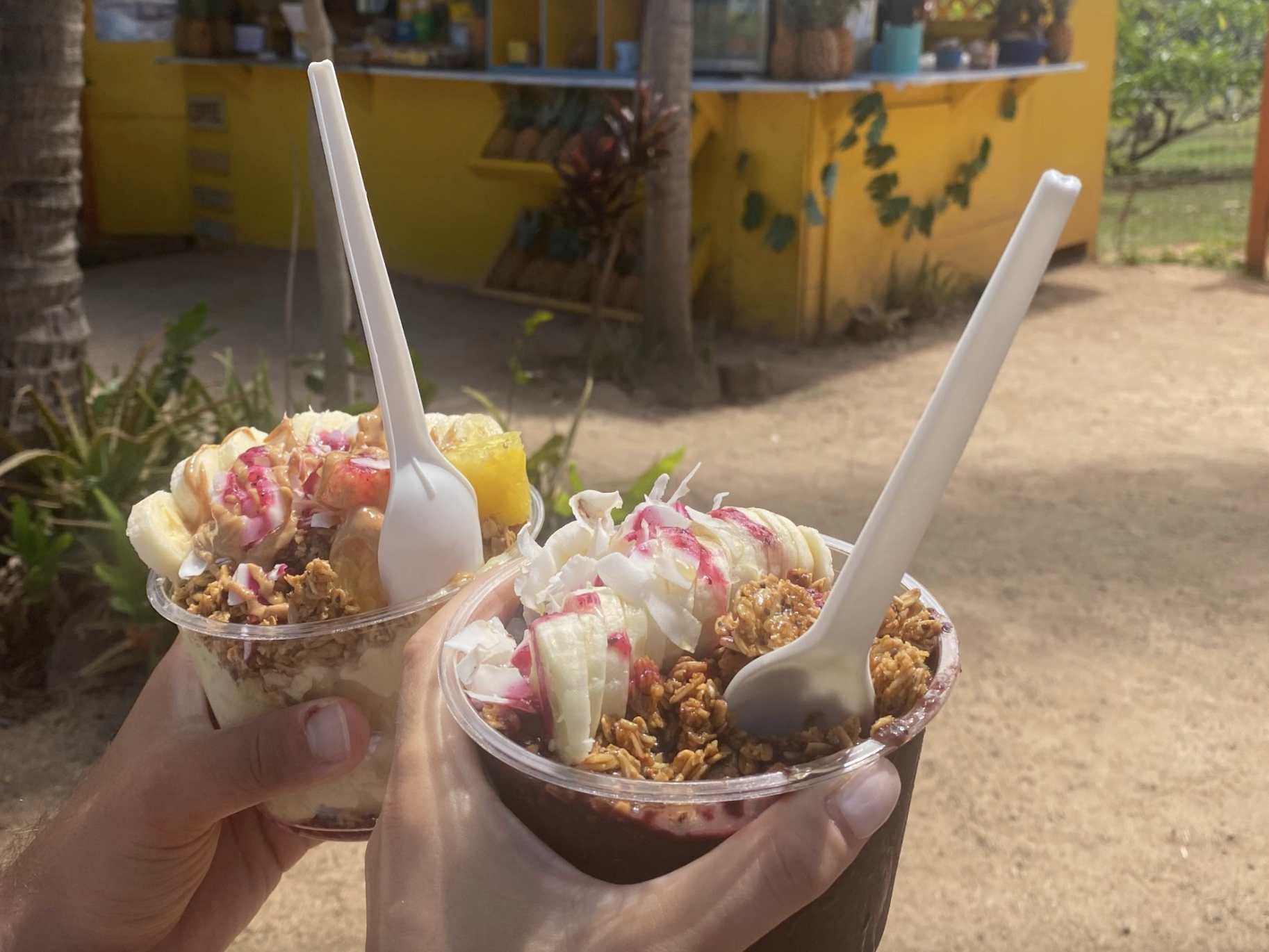 Two acai bowls with white spoons in them and a yellow beach shack in the background.
