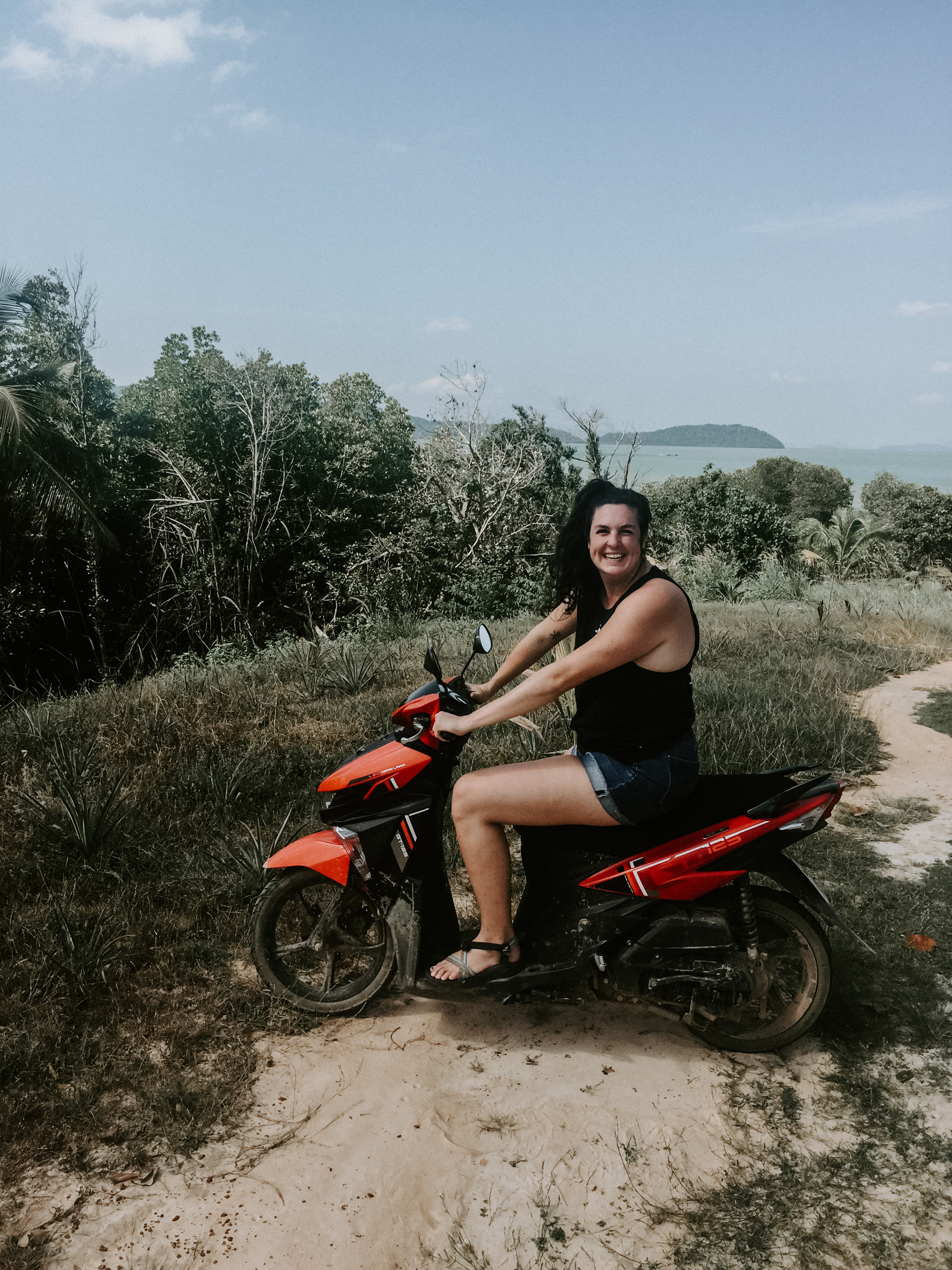 Travel advisor Rachel in a black tank top and shorts riding a red motorbike along a trail with the ocean in view in the distance