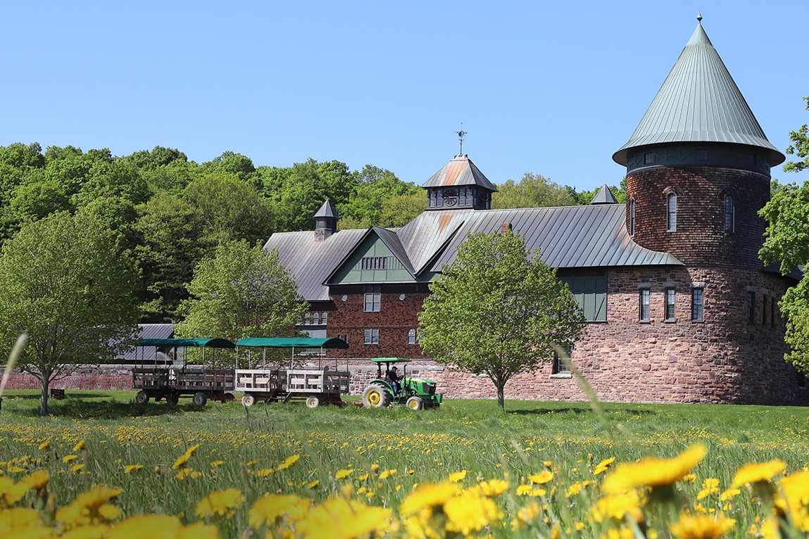 A view of a brick farmhouse with a grassy meadow, trees and wooden carts in the surrounding areas.