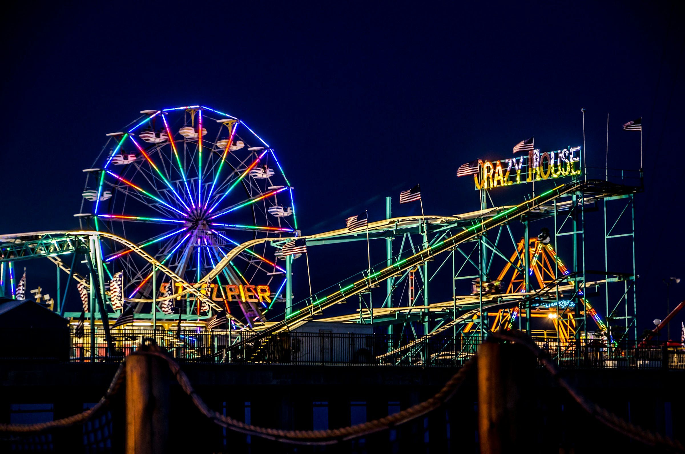 A view of a vibrant neon amusement park at nighttime with rollercoasters and a ferris wheel.