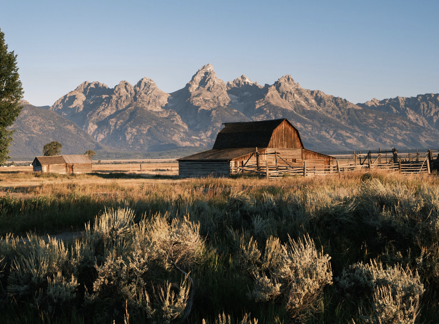 A wooden lodge house in a green field with a lake and rugged mountain terrain in the background.