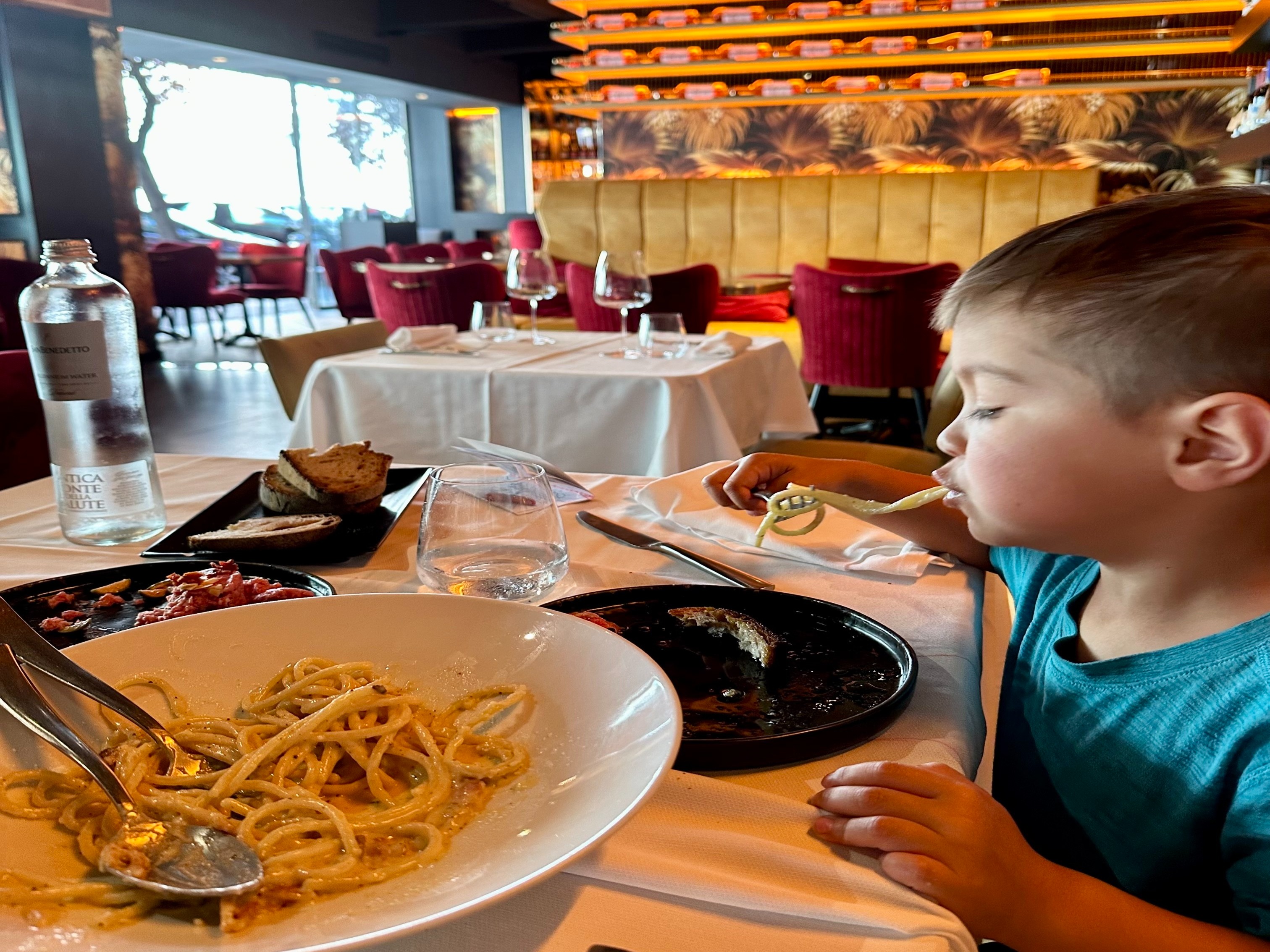 A child eating spaghetti at a restaurant with white tablecloths and red chairs.