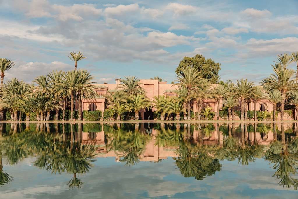 The pink clay edifice of Amanjena stands across a quaint, mirror-flat water feature with palms dotting the shoreline