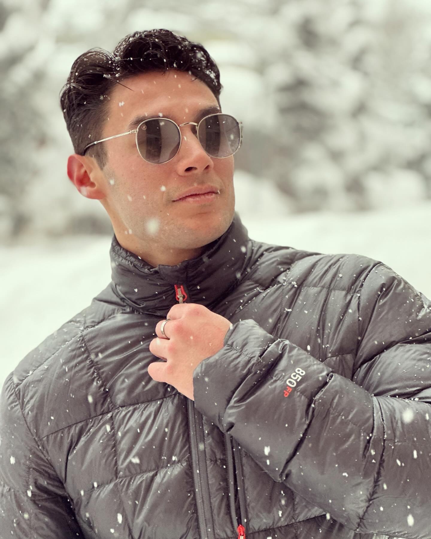 A man tugging on his coat zipper and wearing sunglasses while posing for a photo outside as it snows.