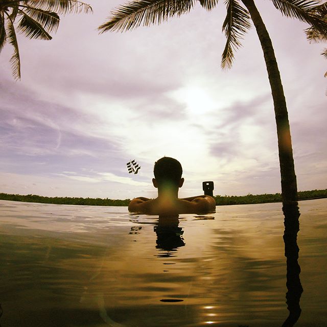 A man relaxing in a swimming pool beneath a cloudy, but sunny sky and palm trees.