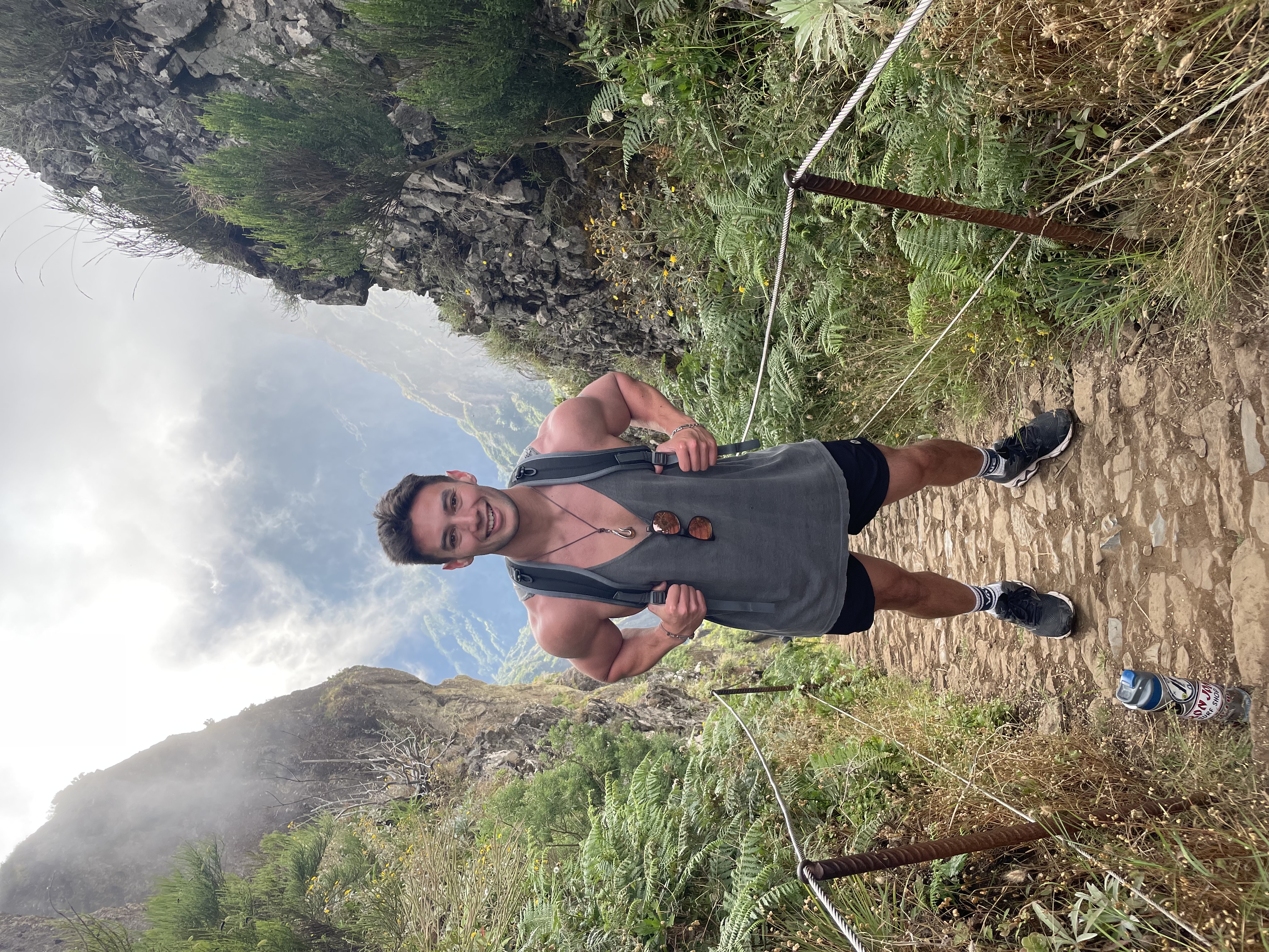 A man posing for a photo in the middle of a hiking trail. There is brush, large rocky cliffs and fog in the surrounding areas.