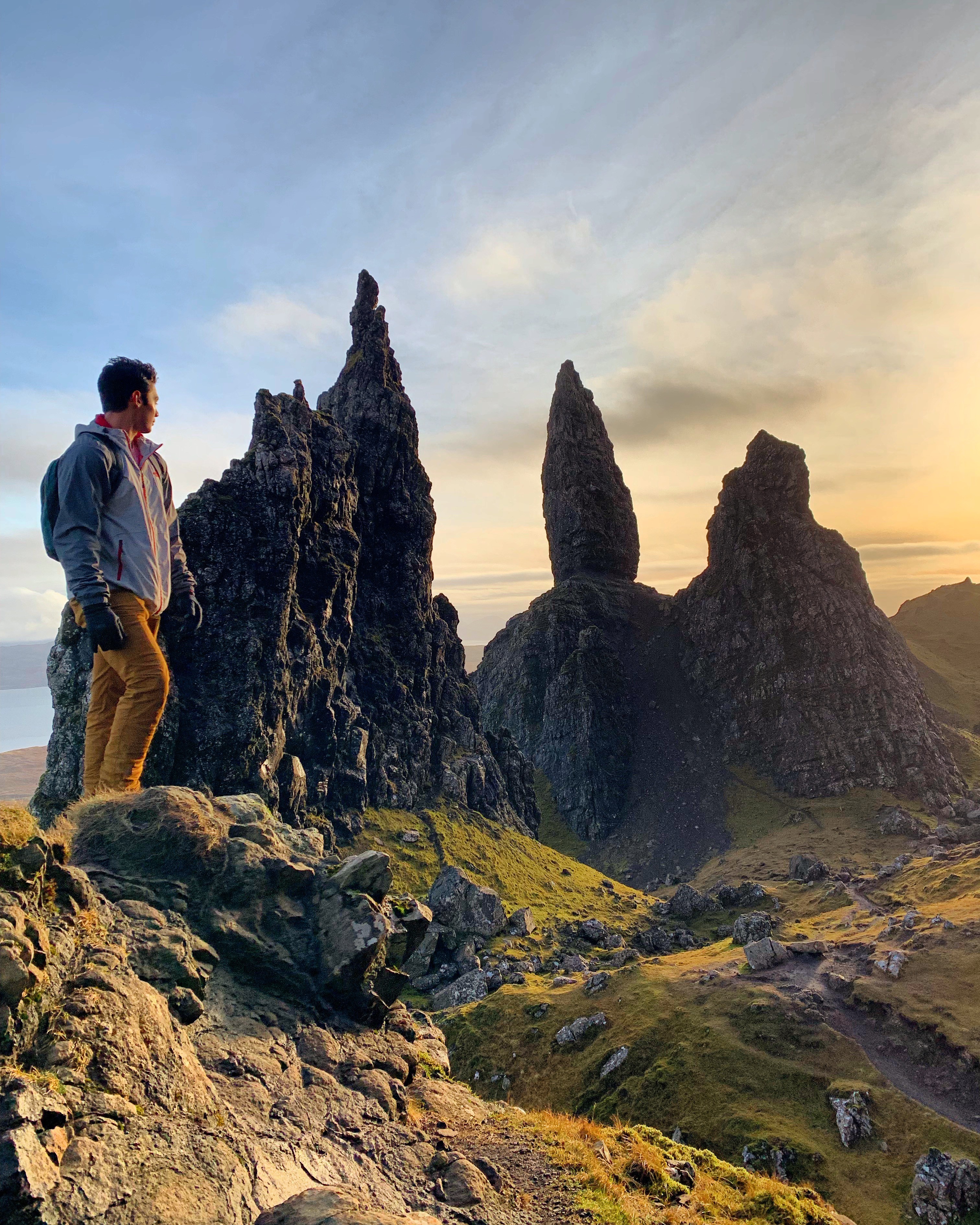 A man looking at a group of large rock formations outside. There is a golden sunset in the distance.