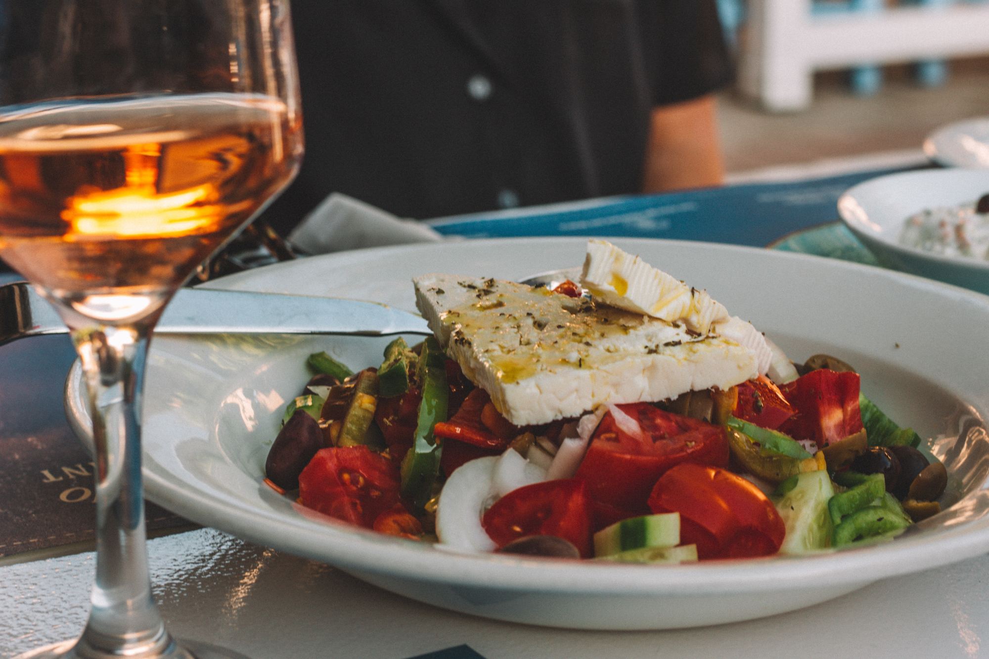 Greek salad on a white plate next to a glass of wine.
