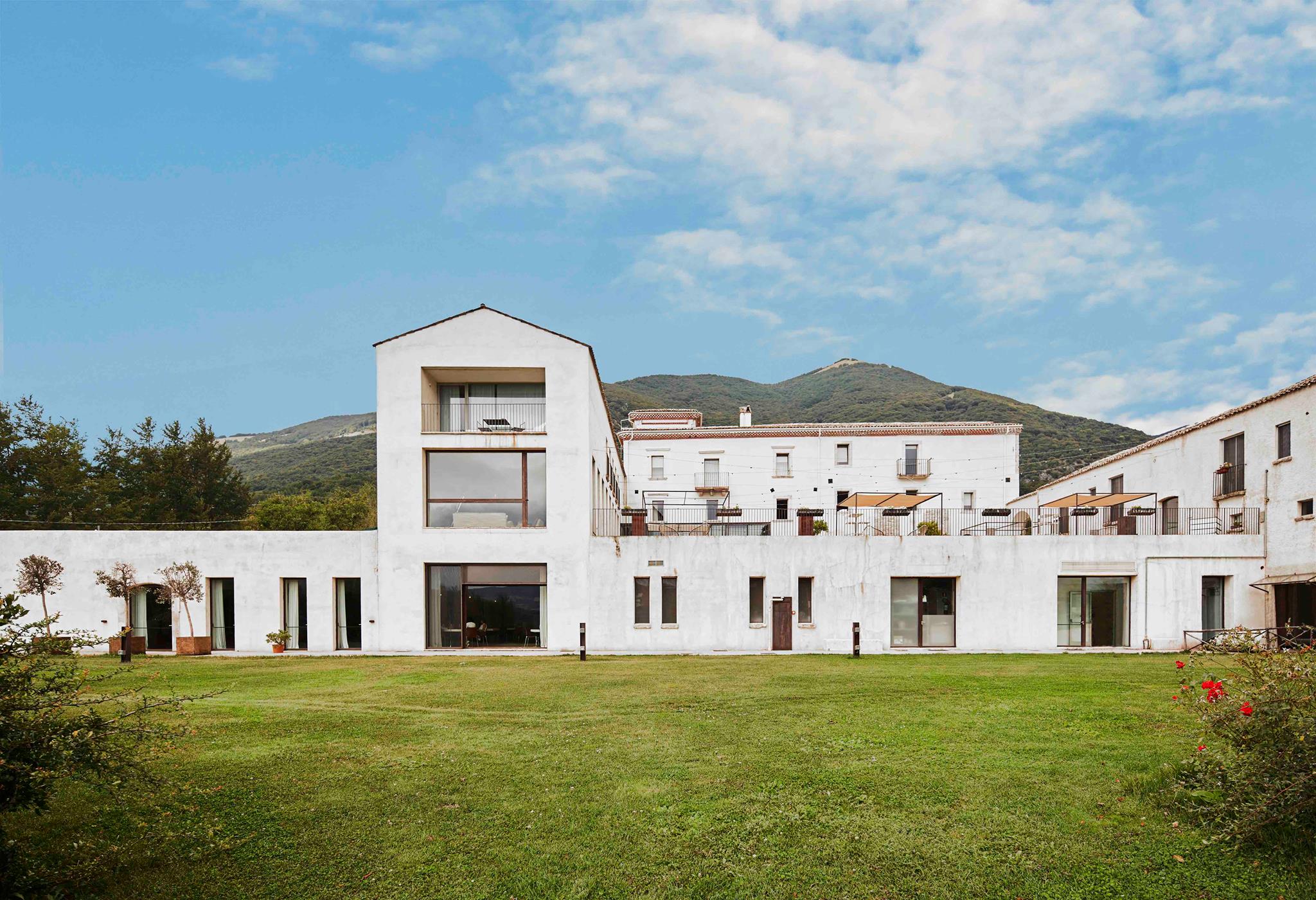 A white villa next to a grassy lawn. There is also a mountain in the background, beneath a cloudy blue sky.