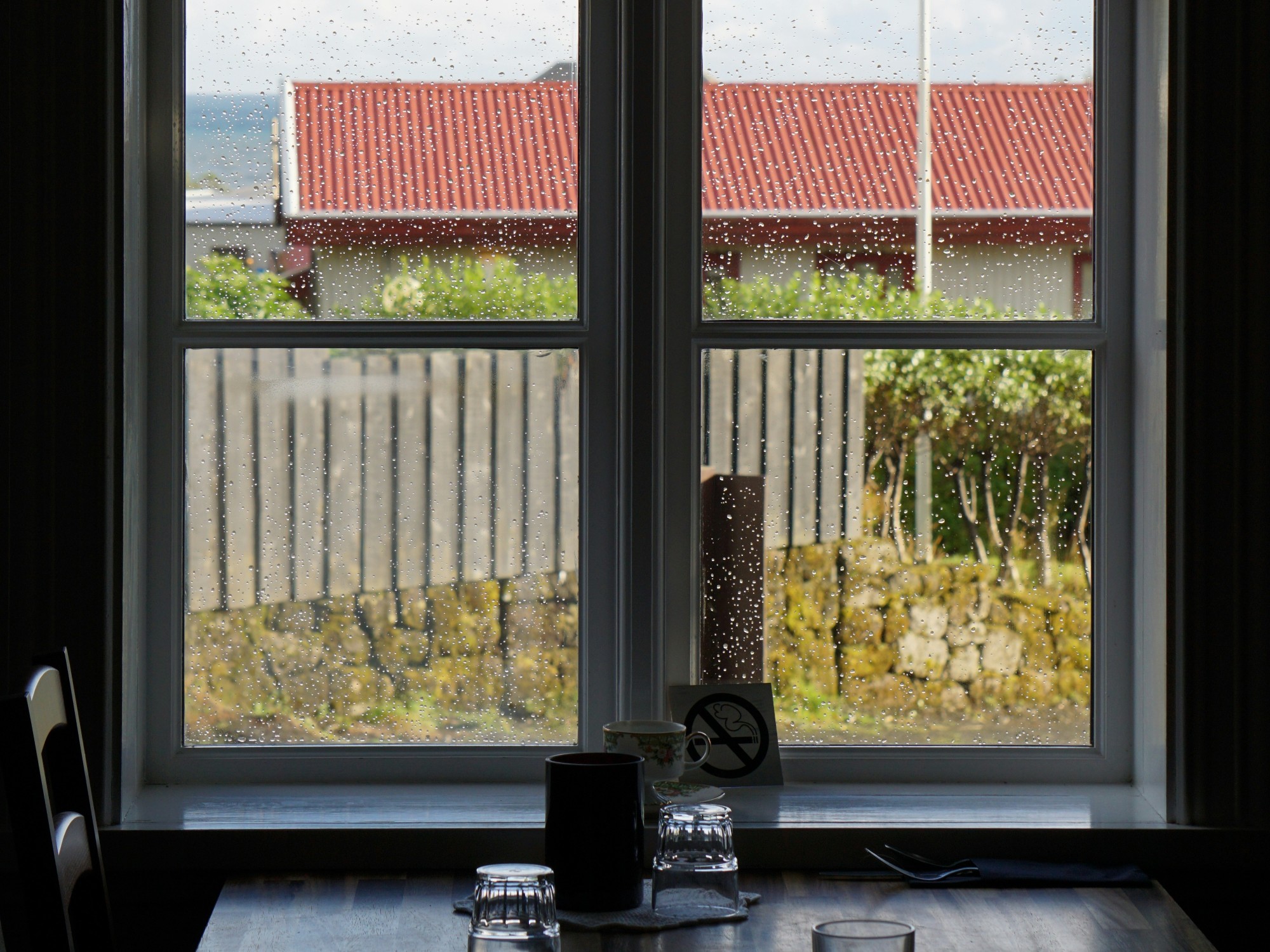 A view of a wooden dining table with glassware placed on top of it next a window with rain droplets on the glass. There is a fence and building with a red roof in the outside view.