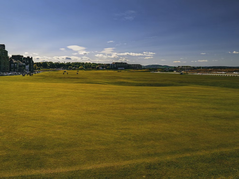 A view of a golf course under a vast blue sky with scattered clouds in the distance.