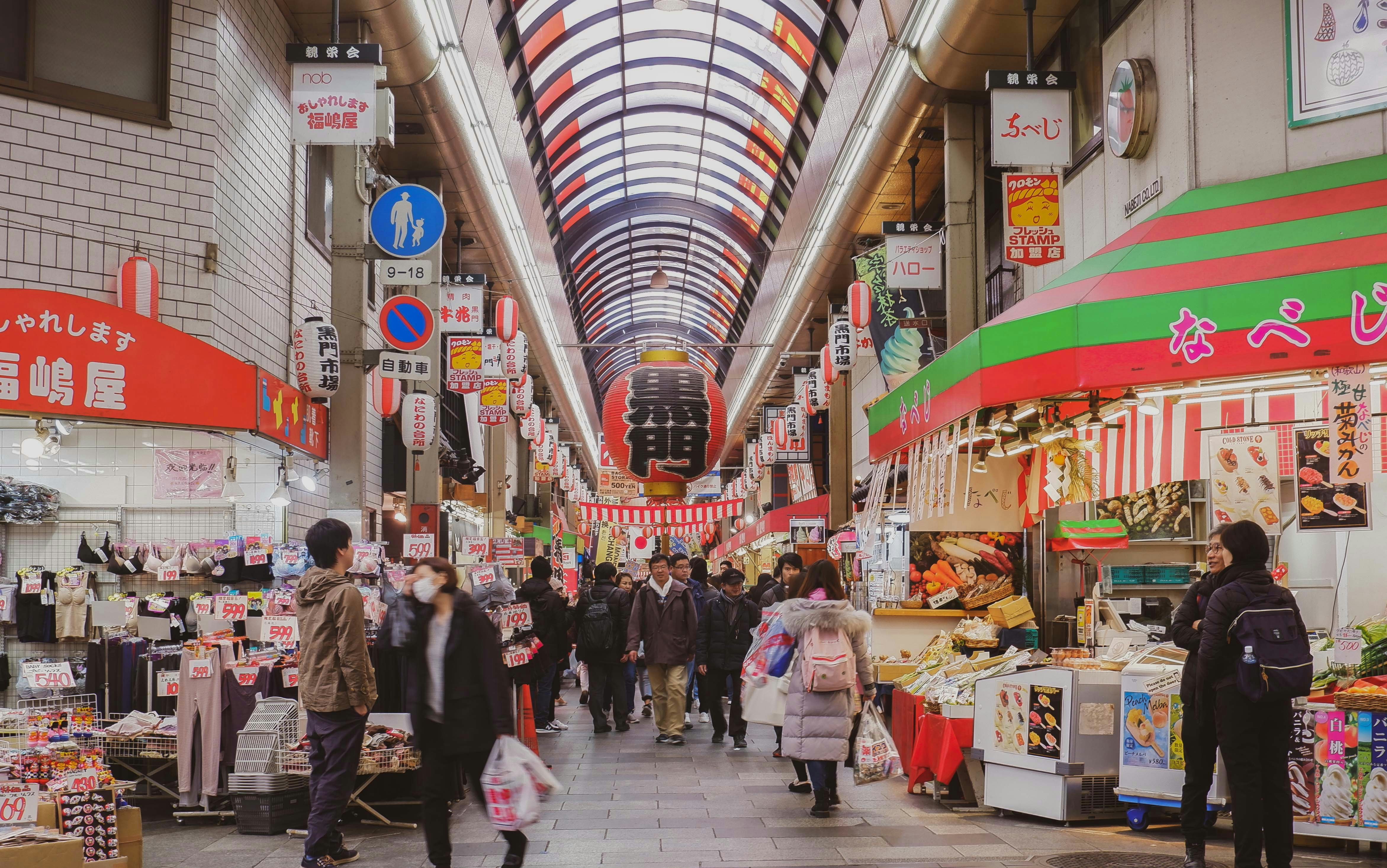 A picture of people walking in a market with different food stalls during the daytime.