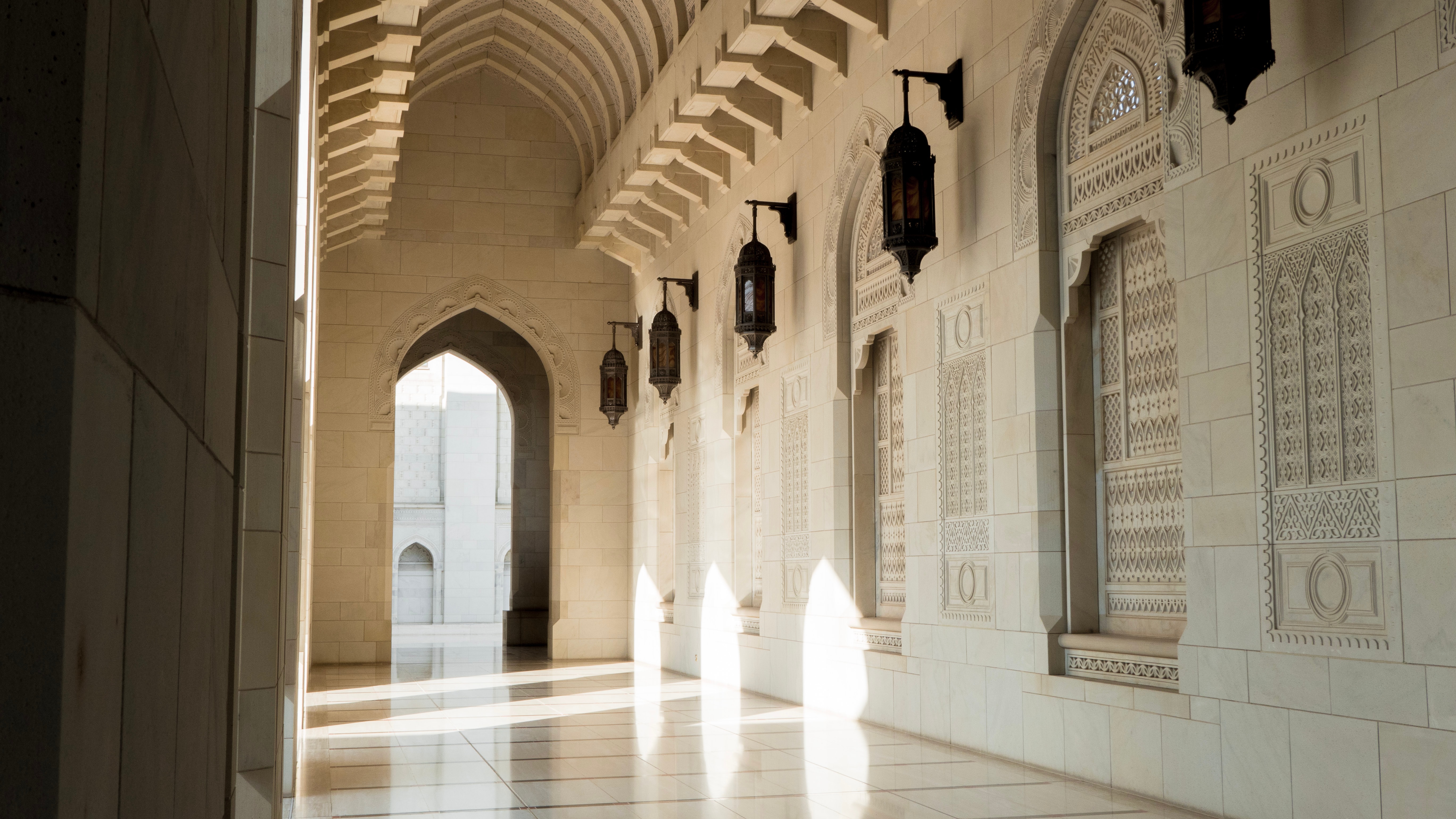 Inside of the Sultan Qaboos Grand Mosque in Oman, complete with cream stone work, beautiful arches and metal lanterns. The sun is shining in through a window against the wall on the right side.