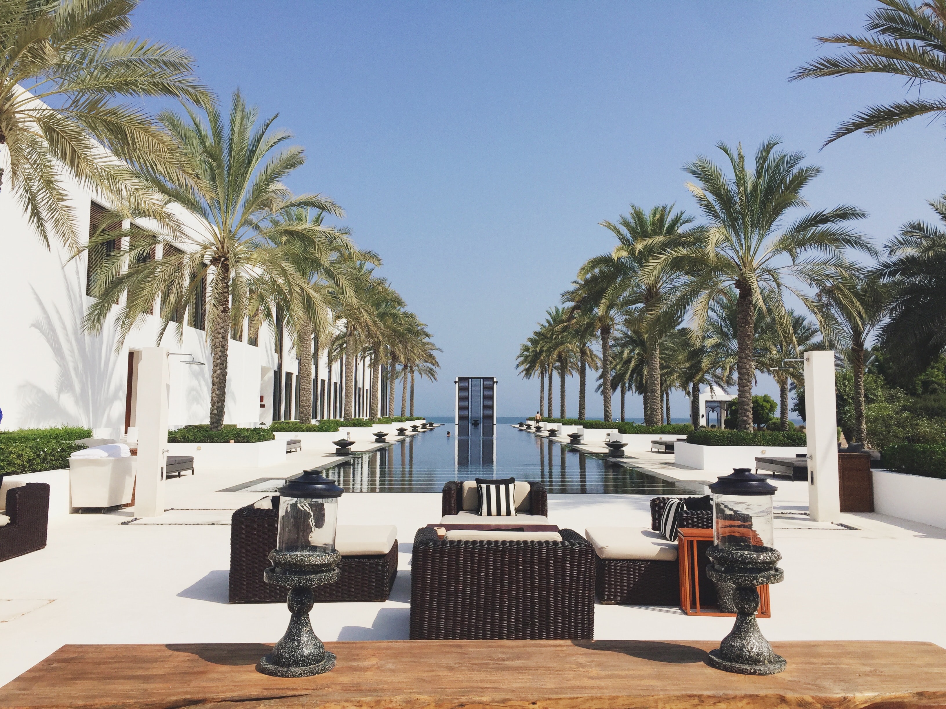 A view of a hotel pool deck with wicker furniture, a wood paneled deck, palm trees, black tables and a luxurious rectangular swimming pool down the middle.