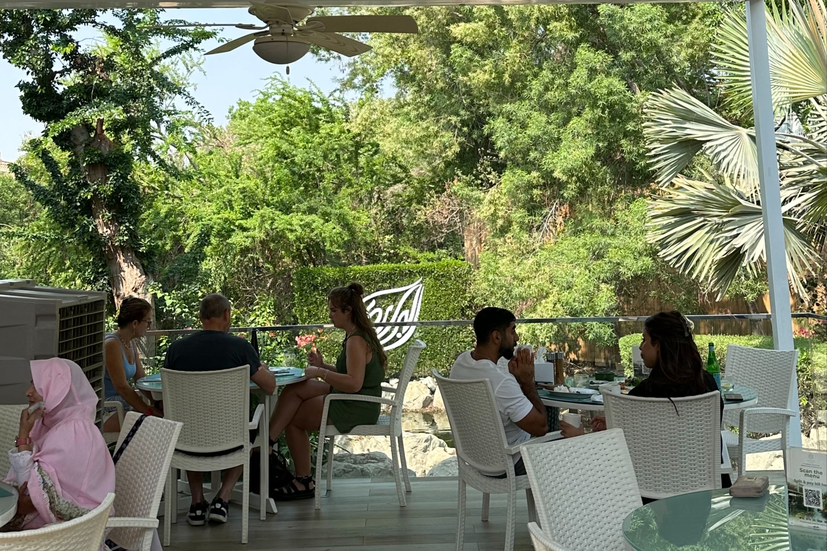 A patio full of white chairs and tables with people seated and dining. There is a lush, green forest full of trees in the background.