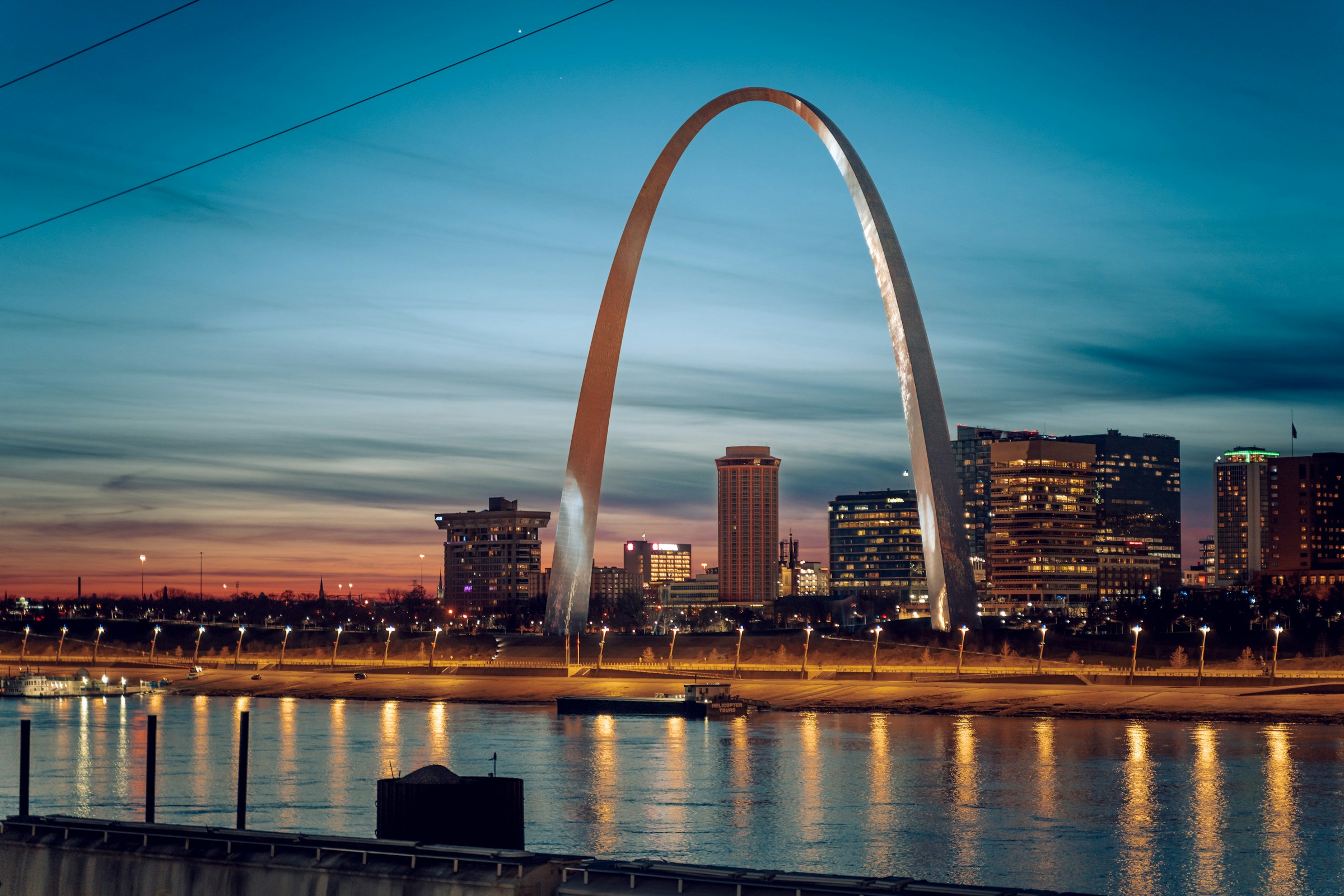 A view of the river and large silver arch in front of the city skyline at night.