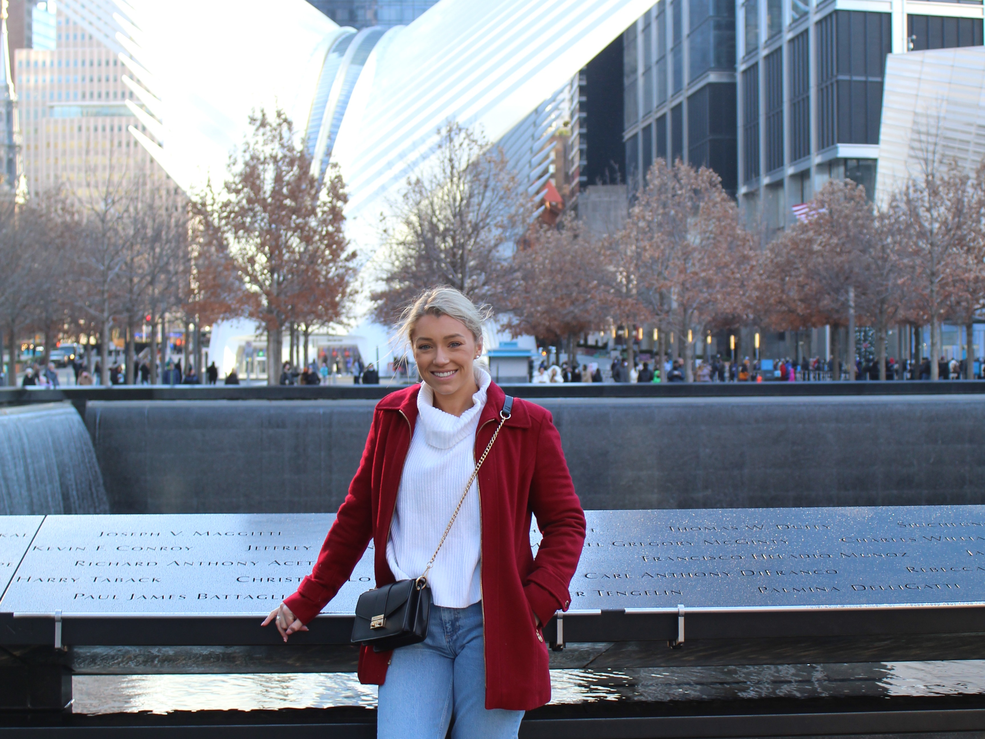 A woman wearing a white top and red coat in front of the 9/11 memorial fountain. There are trees and large buildings in the background.