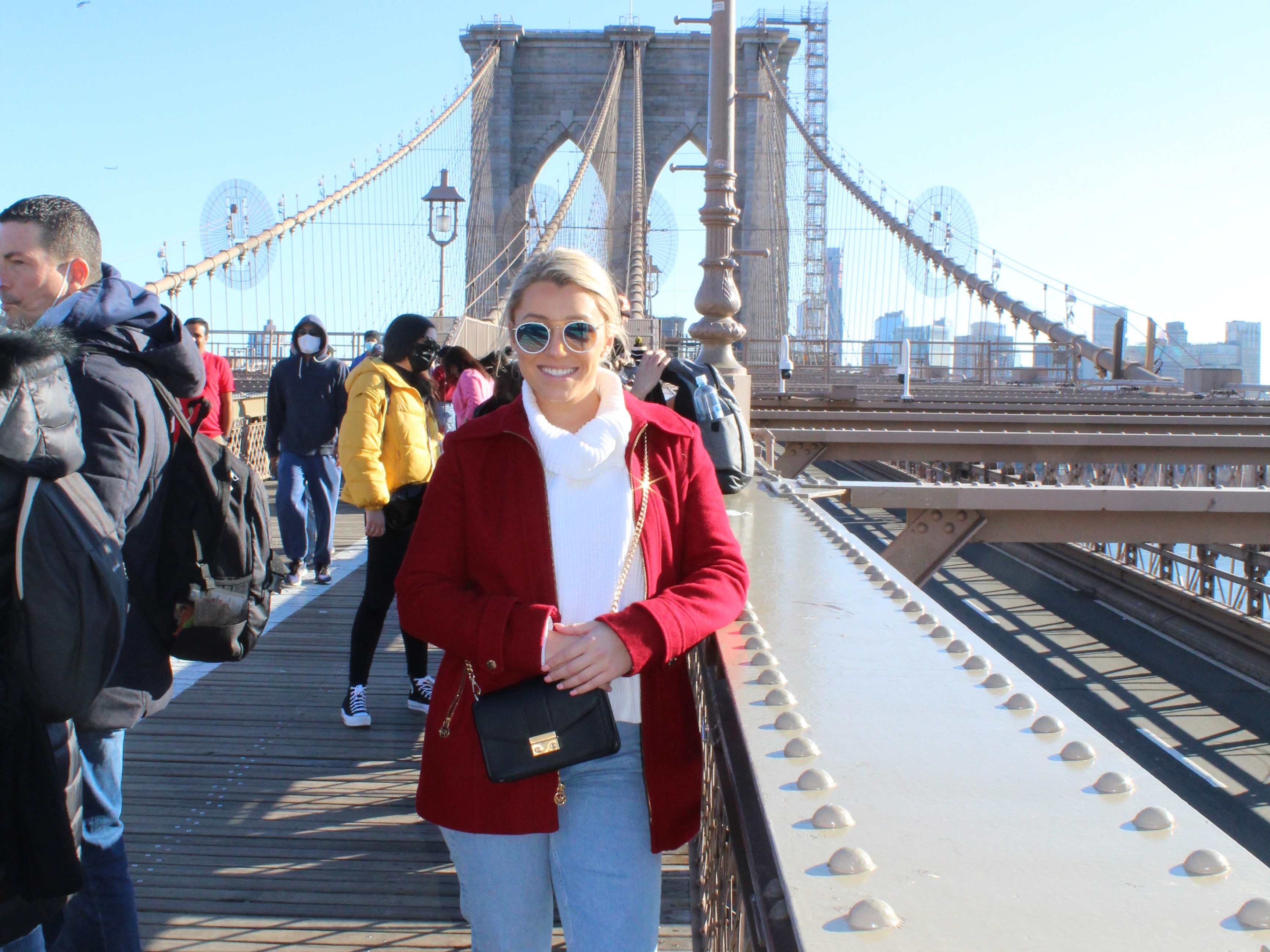 A woman wearing a white top and red jacket posing on the Brooklyn Bridge with many people walking around in the surrounding areas on a bright, sunny day.