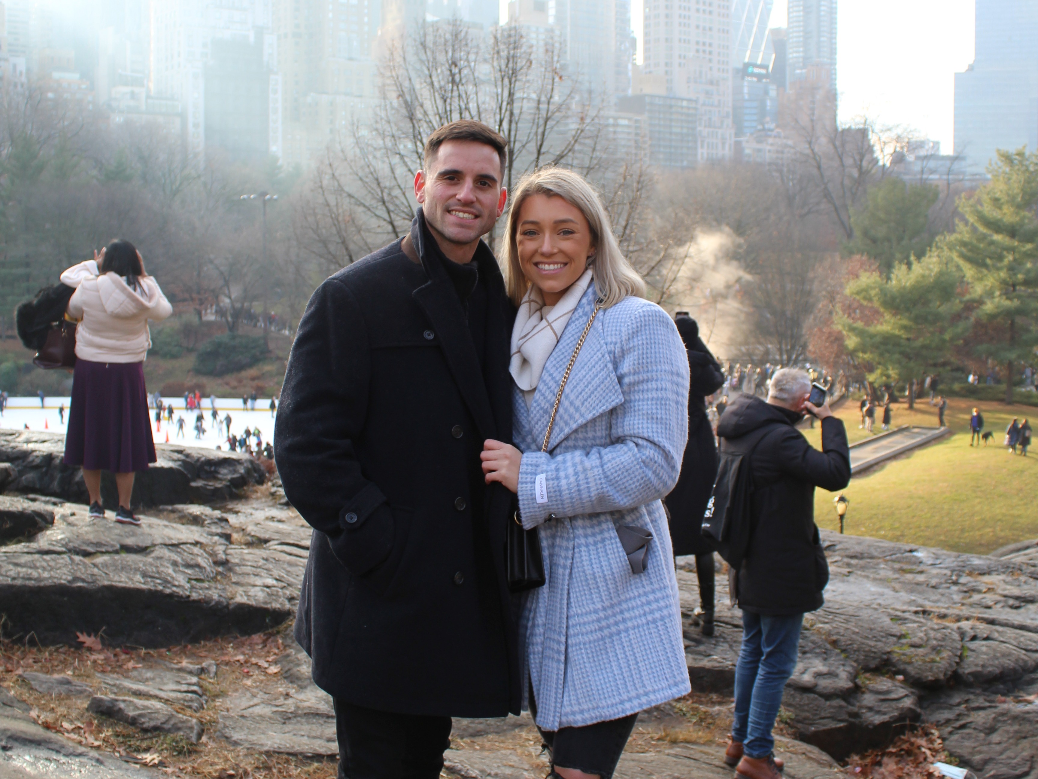 A couple posing for a photo in Central Park. There are people in the background, as well as trees, grass and tall buildings.