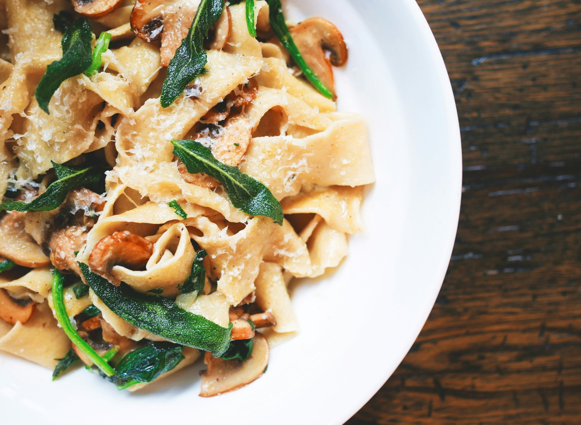 A white plate full of pasta and greens on top of a wooden table.