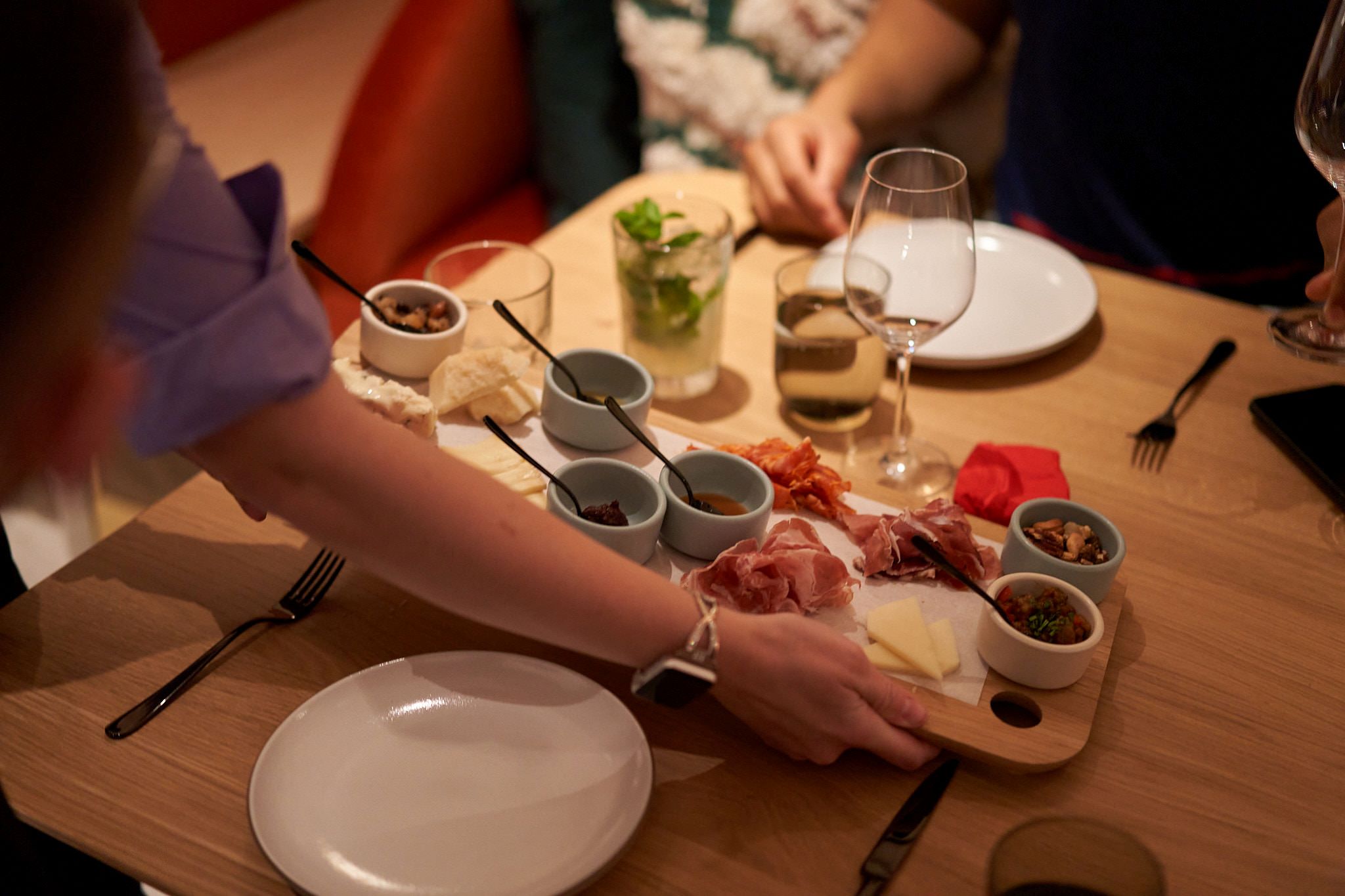 A wooden table full of plates of food, silverware and wine glasses. There is a person holding a wooden charcuterie board with various toppings placing it down onto the table.