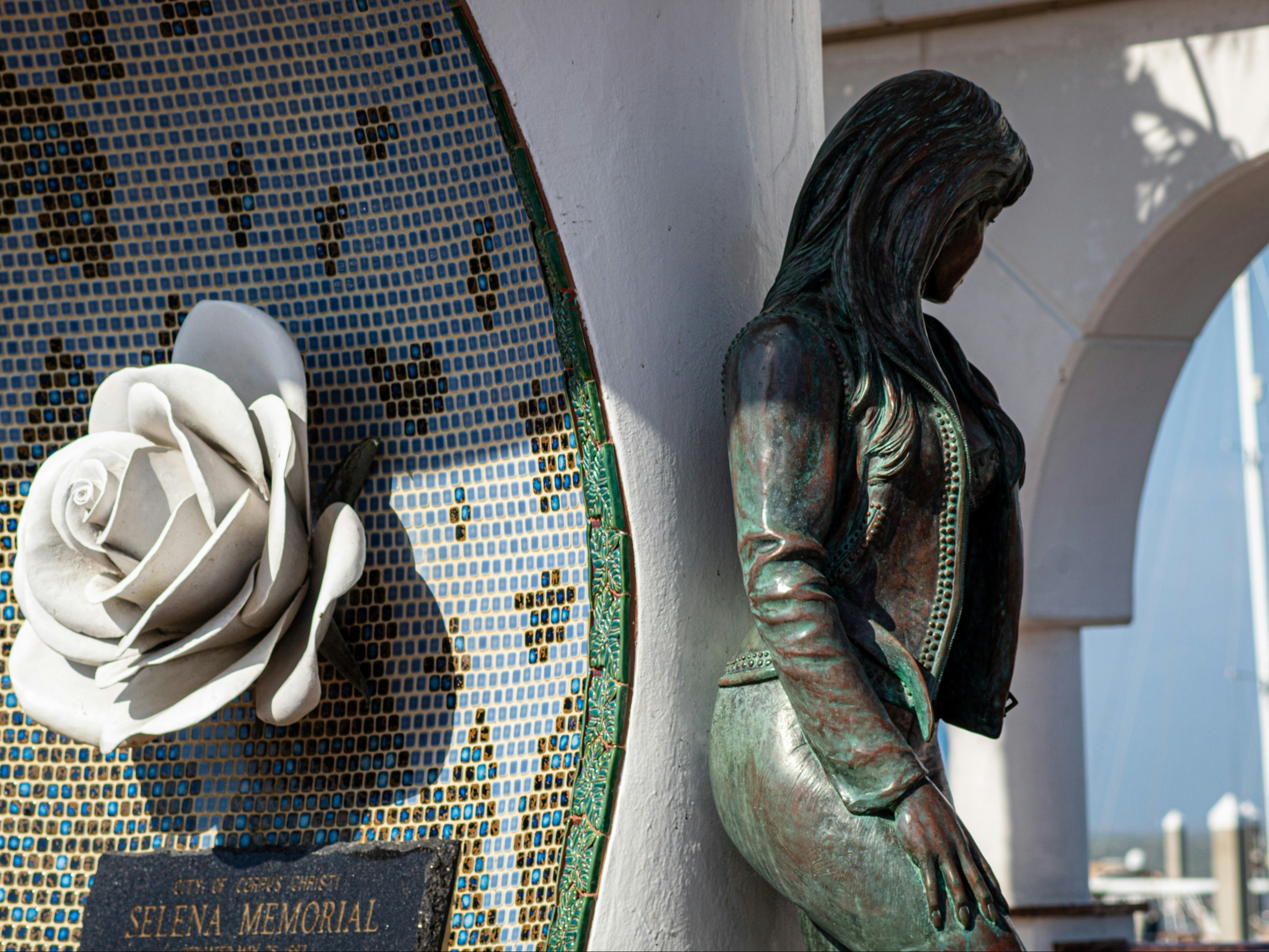 A picture taken in Mirador de la Flor, the Selena Memorial, in Corpus Christi during daytime. It showcases a woman and a large white rose with green and blue tiling.
