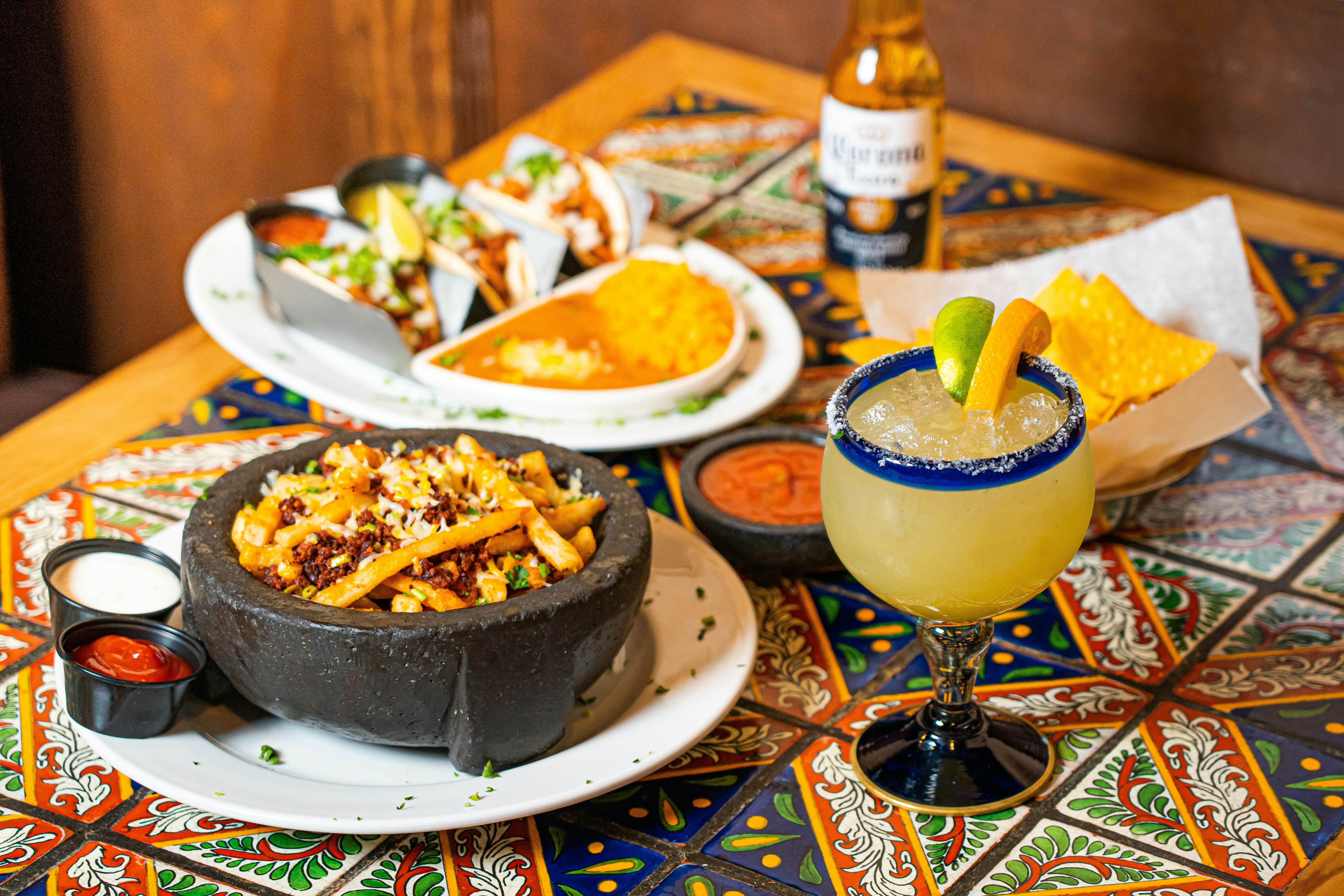 A mosaic tiled table with various plates of latin cuisine, followed by a margarita and a bottle of beer in the surrounding areas.