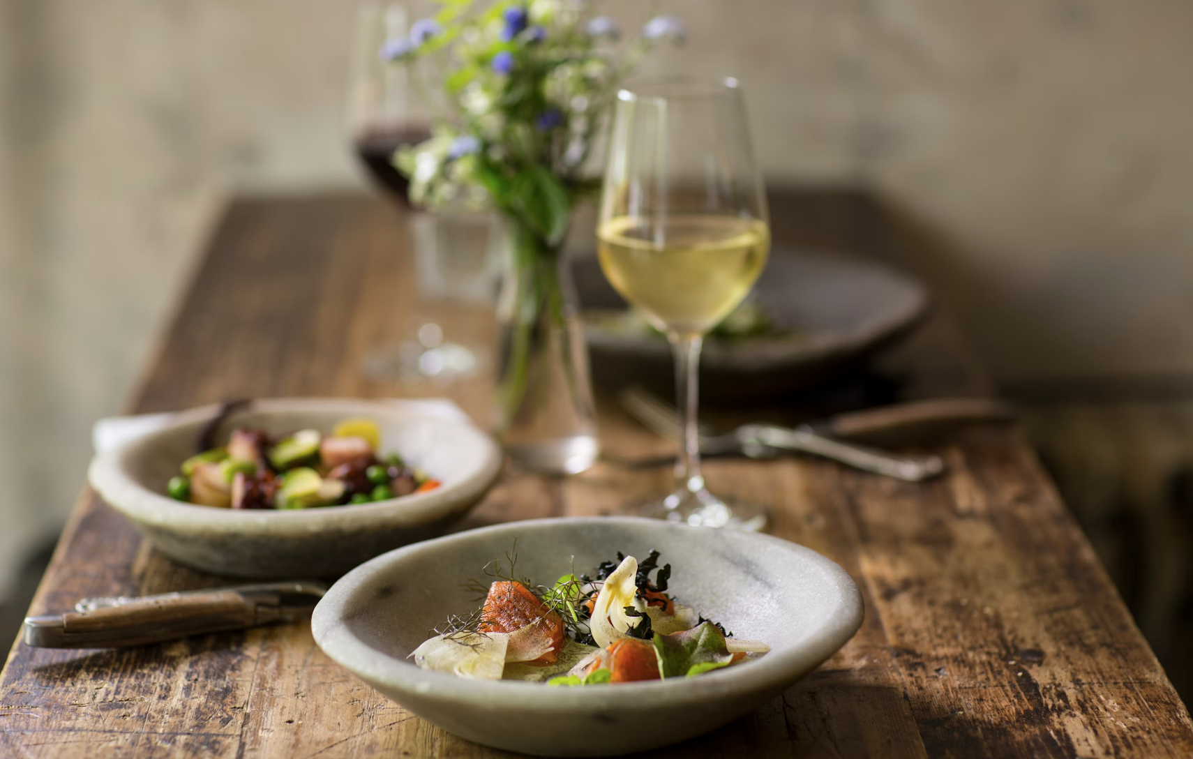 Three plates of colorful food with silverware and wine glasses on top of a wooden table. There is a floral arrangement in the middle of the table as well.