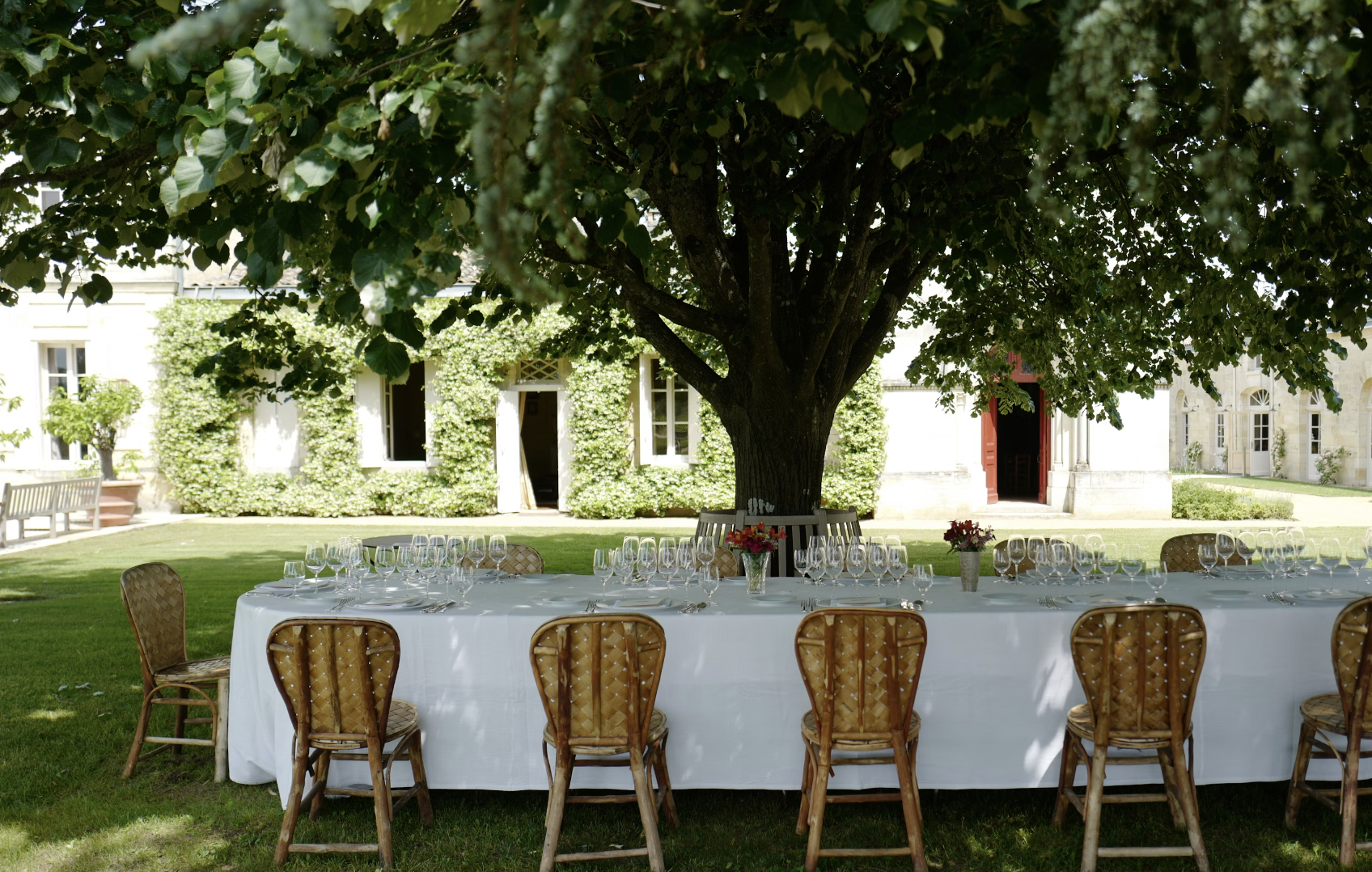 A beautiful outdoor dining table with wicker chairs, a manicured lawn, abundant tree and stone house covered in vines in the background.