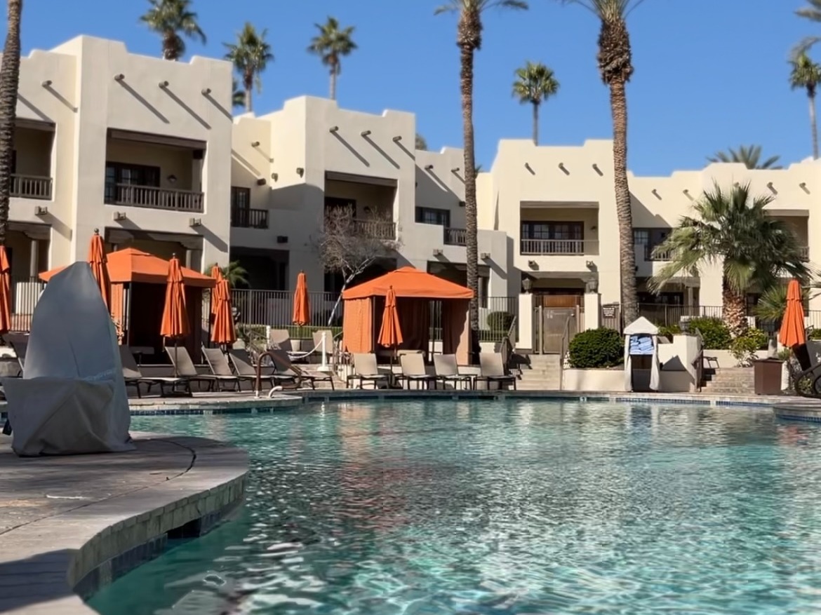 A beautiful view of a hotel swimming pool deck with orange cabanas, lawn chairs, palm trees and stone buildings in the background.