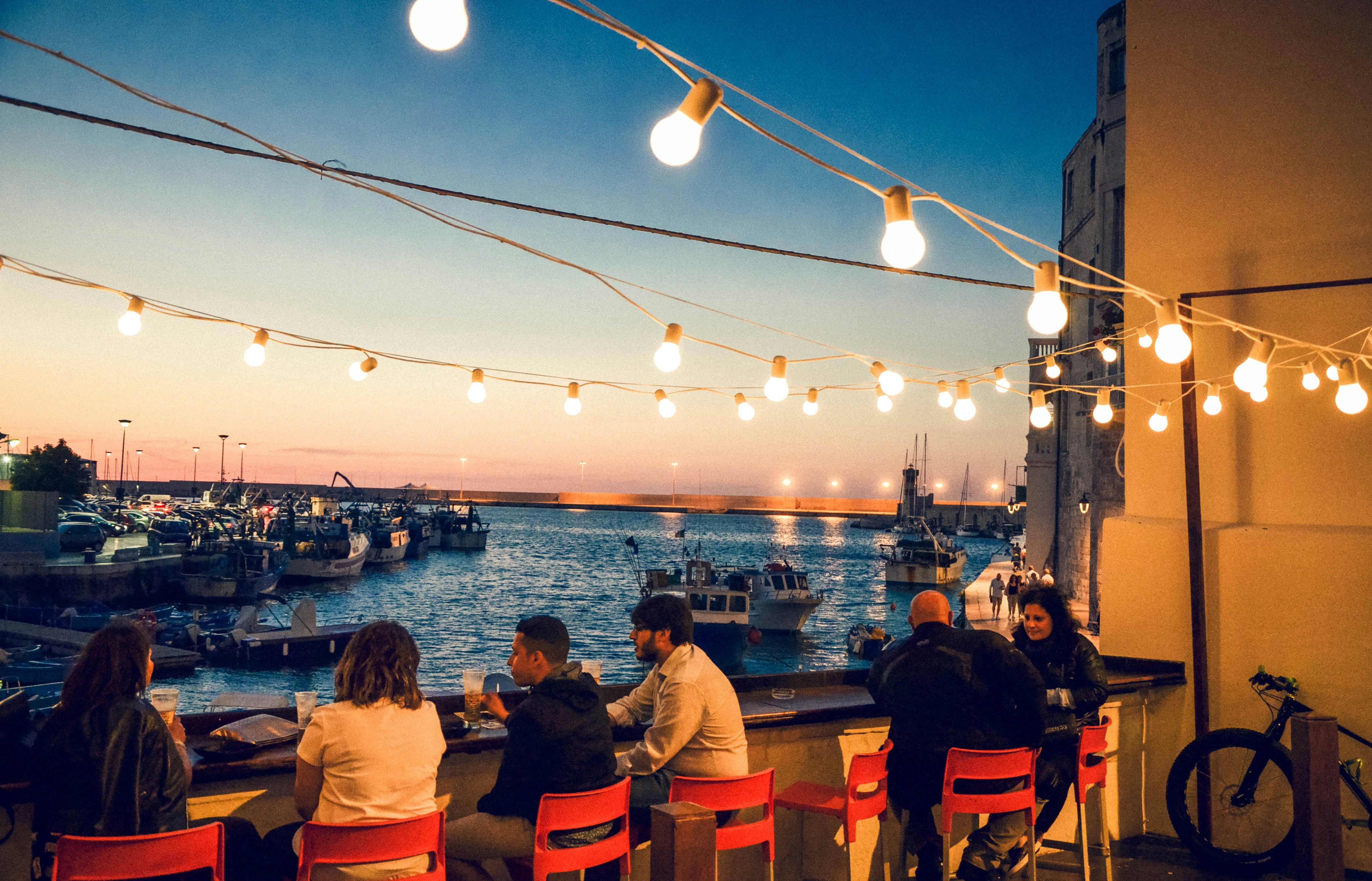 A beautiful view of string lights cascading over people dining at a restaurant outdoors while overlooking a sunset over the harbor.