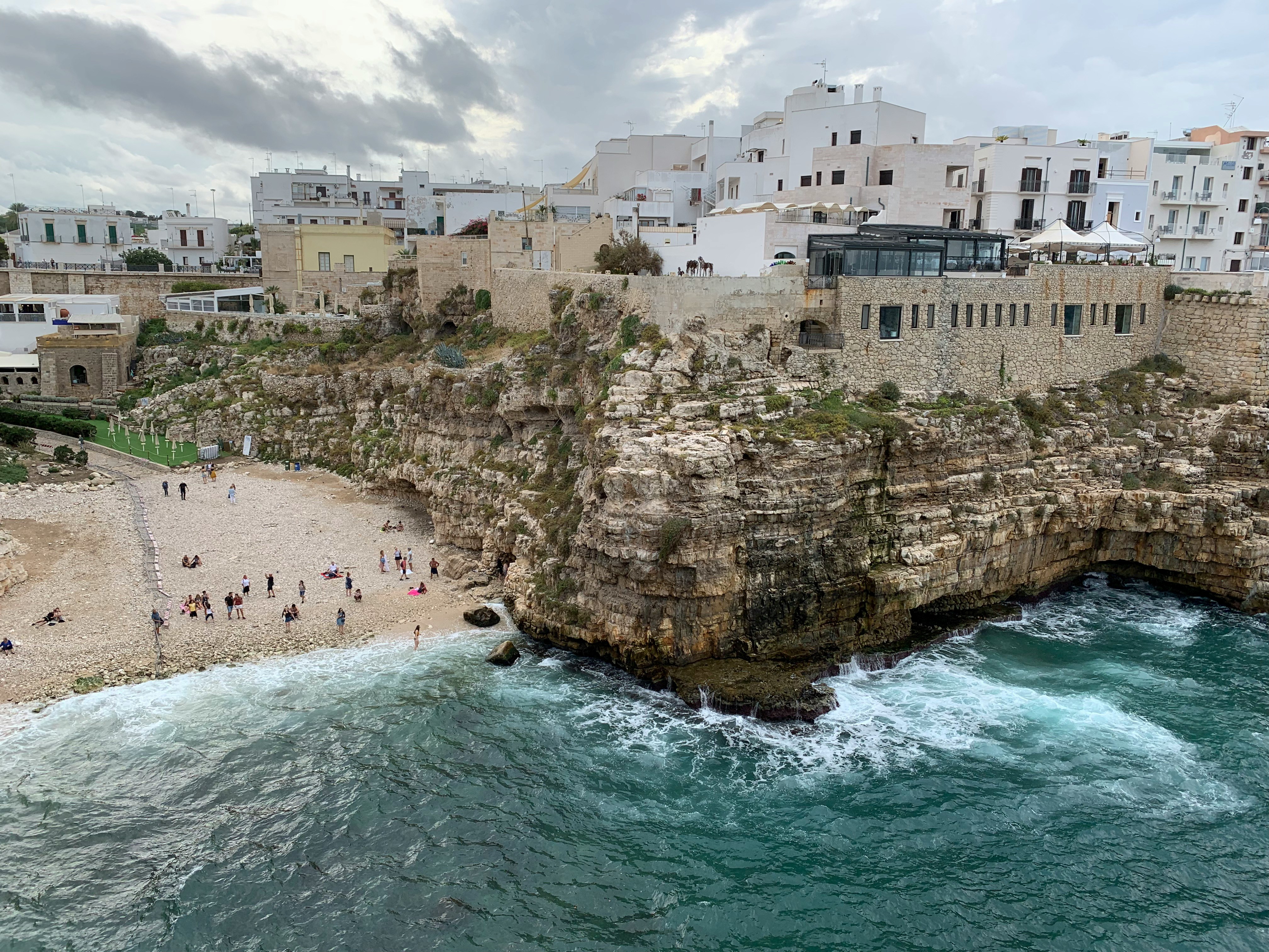 An aerial view of the Polignano a Mare, with a view people swimming, snorkeling and cliff diving amidst the rocky and sandy shoreline and buildings.
