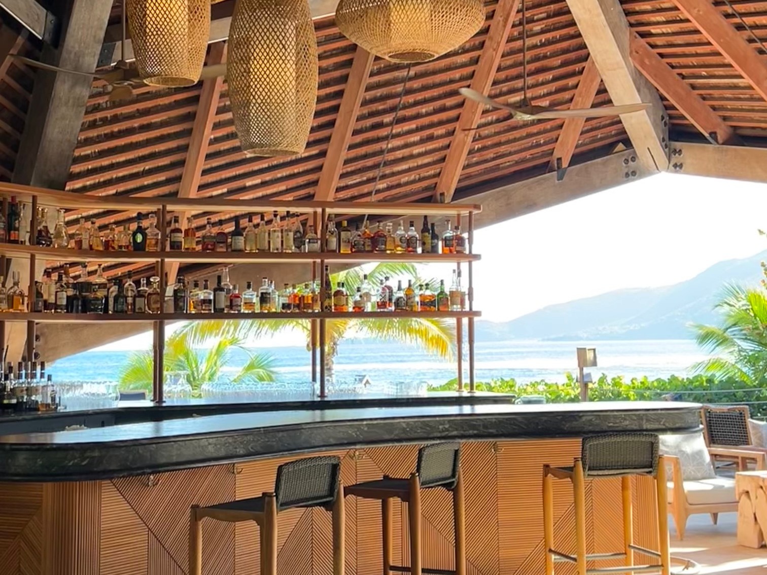 An outdoor bar with alcohol on a shelf, wood paneled ceilings and a view of the mountains and palm trees in the distance.